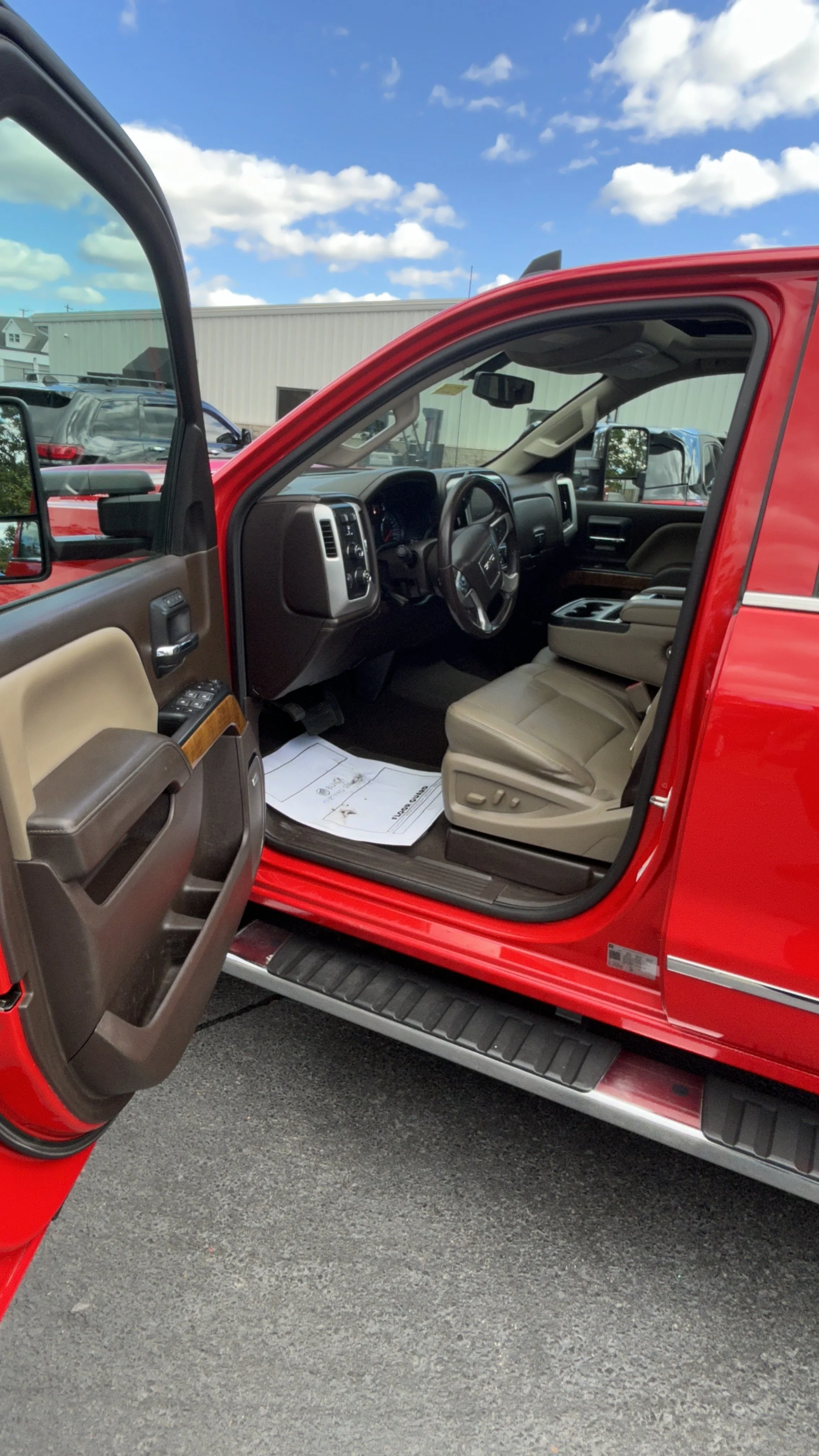 The interior of a red pickup truck, showing the driver's seat, steering wheel, dashboard, and open door with window controls, with other vehicles and a building in the background under a blue sky with clouds.