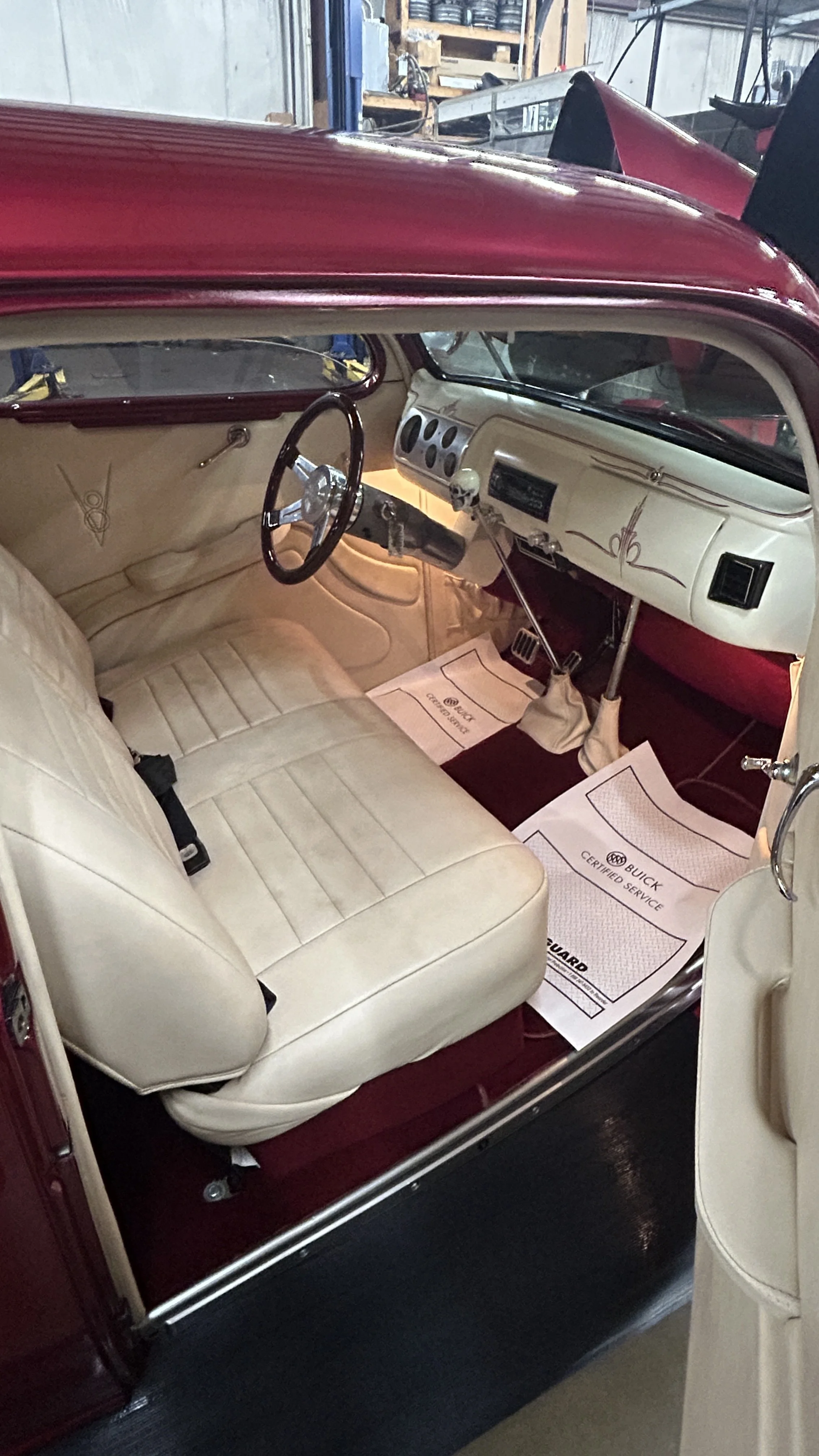 The interior of a vintage car with cream-colored leather seats, a cream dashboard with air vents and gauges, a wooden steering wheel, and floor mats with Buick branding.