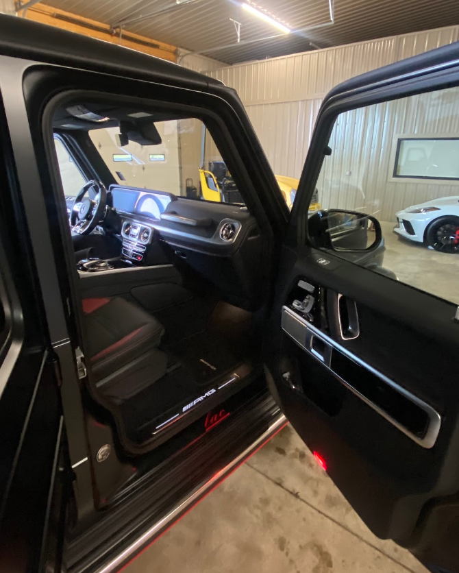 Interior of a black luxury SUV in a garage, with the driver's side door open, revealing the dashboard, steering wheel, and seat, and other cars visible in the background.