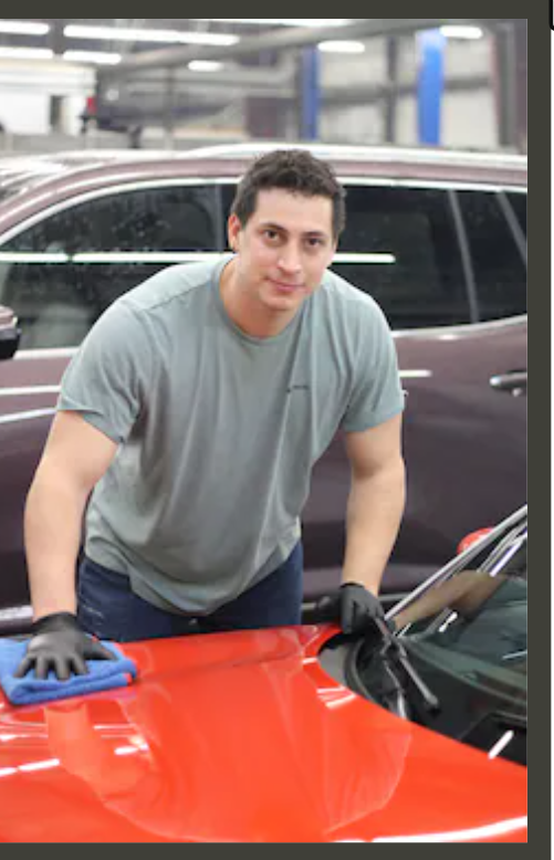 A young man in a gray t-shirt and black gloves cleaning or polishing a red car with a cloth inside a garage or auto shop.