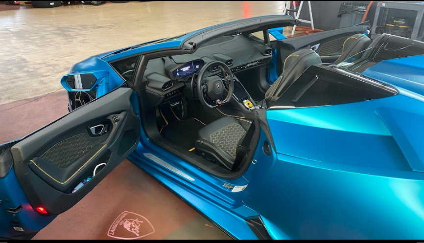 Interior of a blue Lamborghini sports car, showing the open driver’s side door, steering wheel, digital dashboard, and seats.
