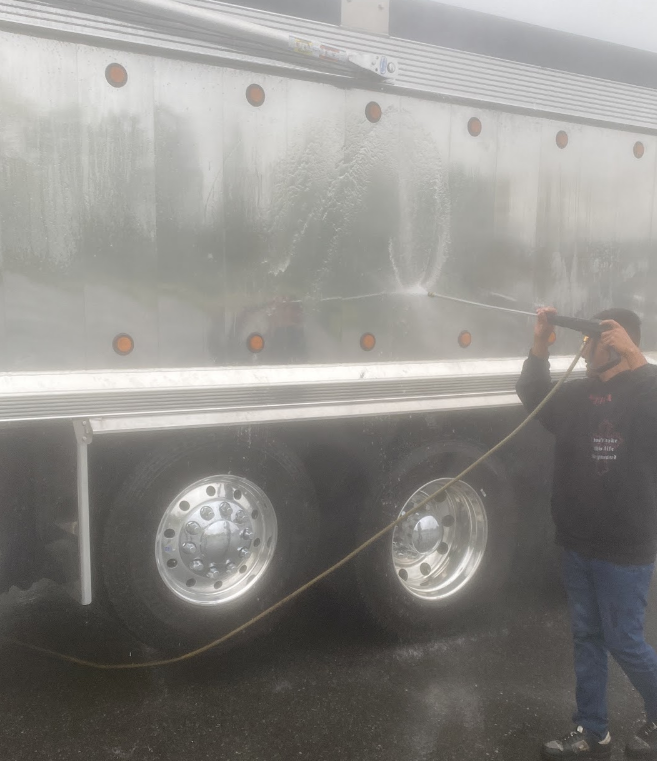 Person using a pressure washer to clean the side of a large silver truck, with wet metal surface and shiny wheels.