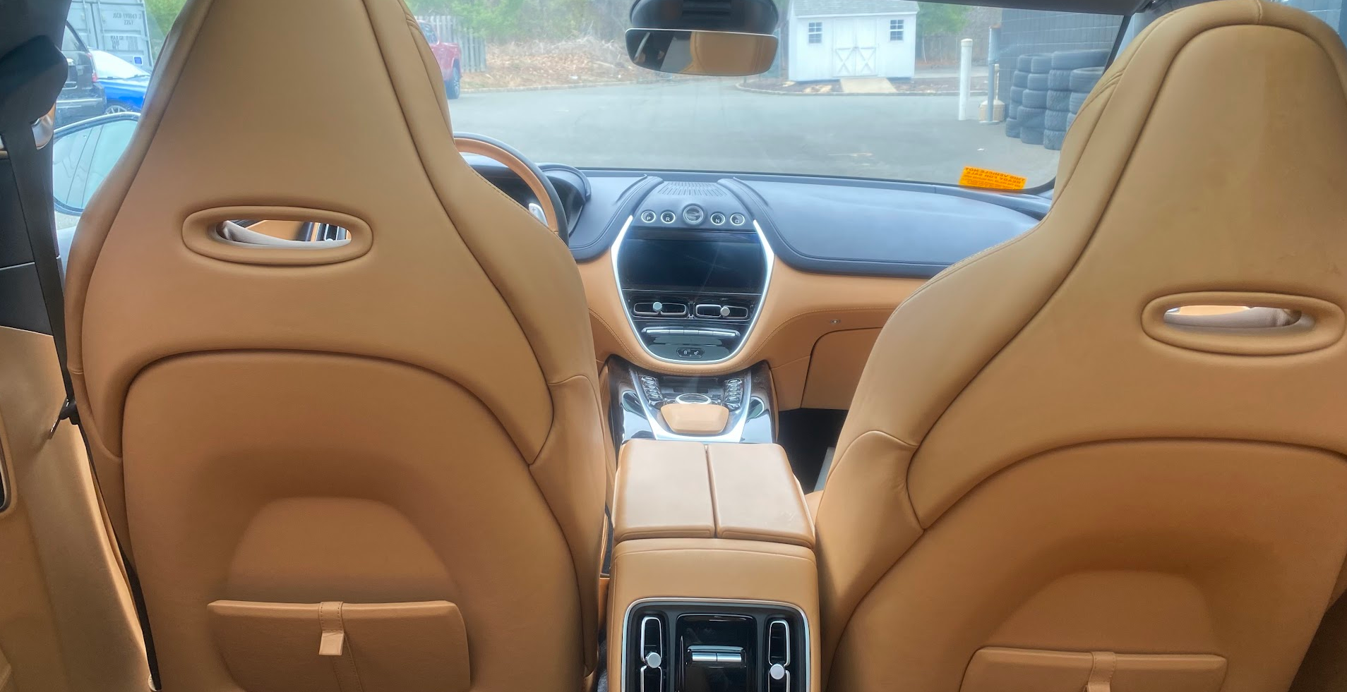 Interior of a luxury car viewed from the back seat, featuring beige leather seats, a central console with controls, and a dashboard with a screen and air vents. Outside the windshield, a parking lot with stacked tires, a building, and other vehicles 