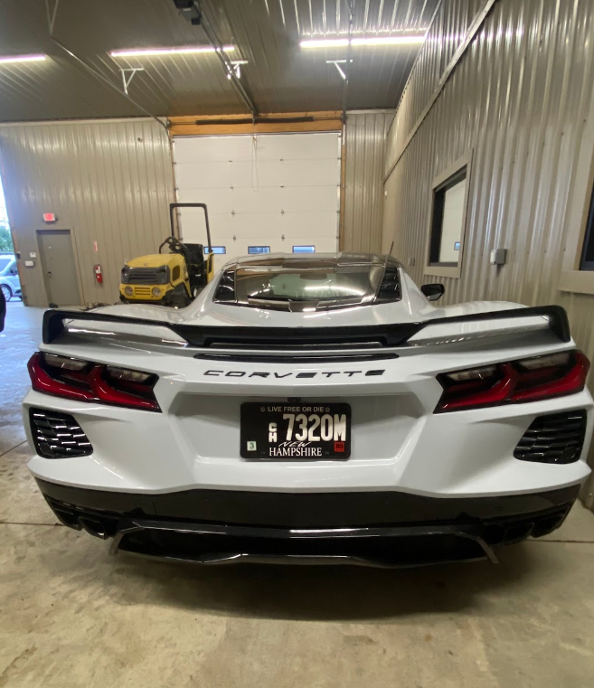 White Chevrolet Corvette inside a garage with a black spoiler, license plate from Hampshire, and a yellow machine in the background.