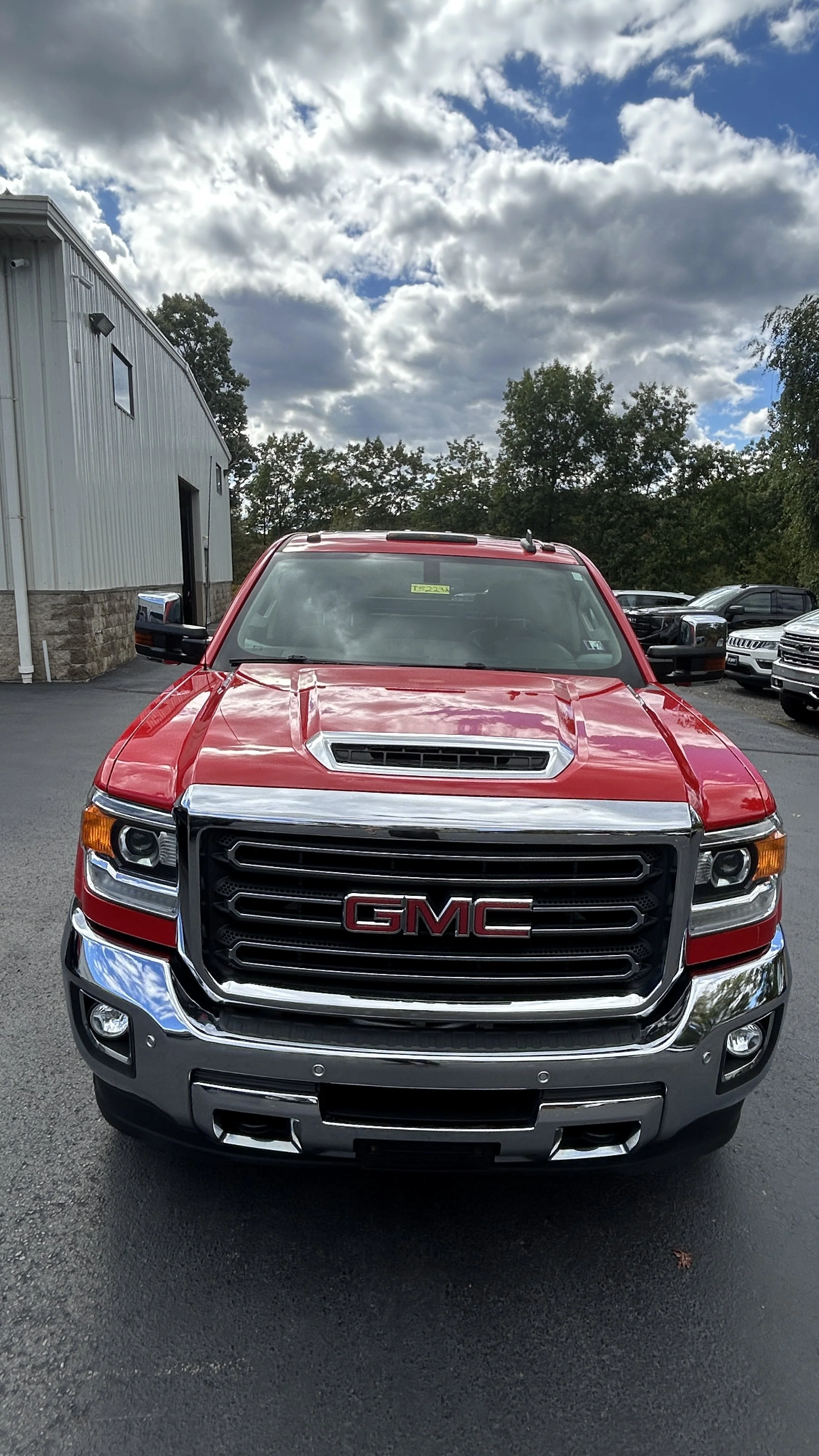 Front view of a red GMC pickup truck parked on a paved lot, with a building to its left and several other vehicles in the background, under a partly cloudy sky.