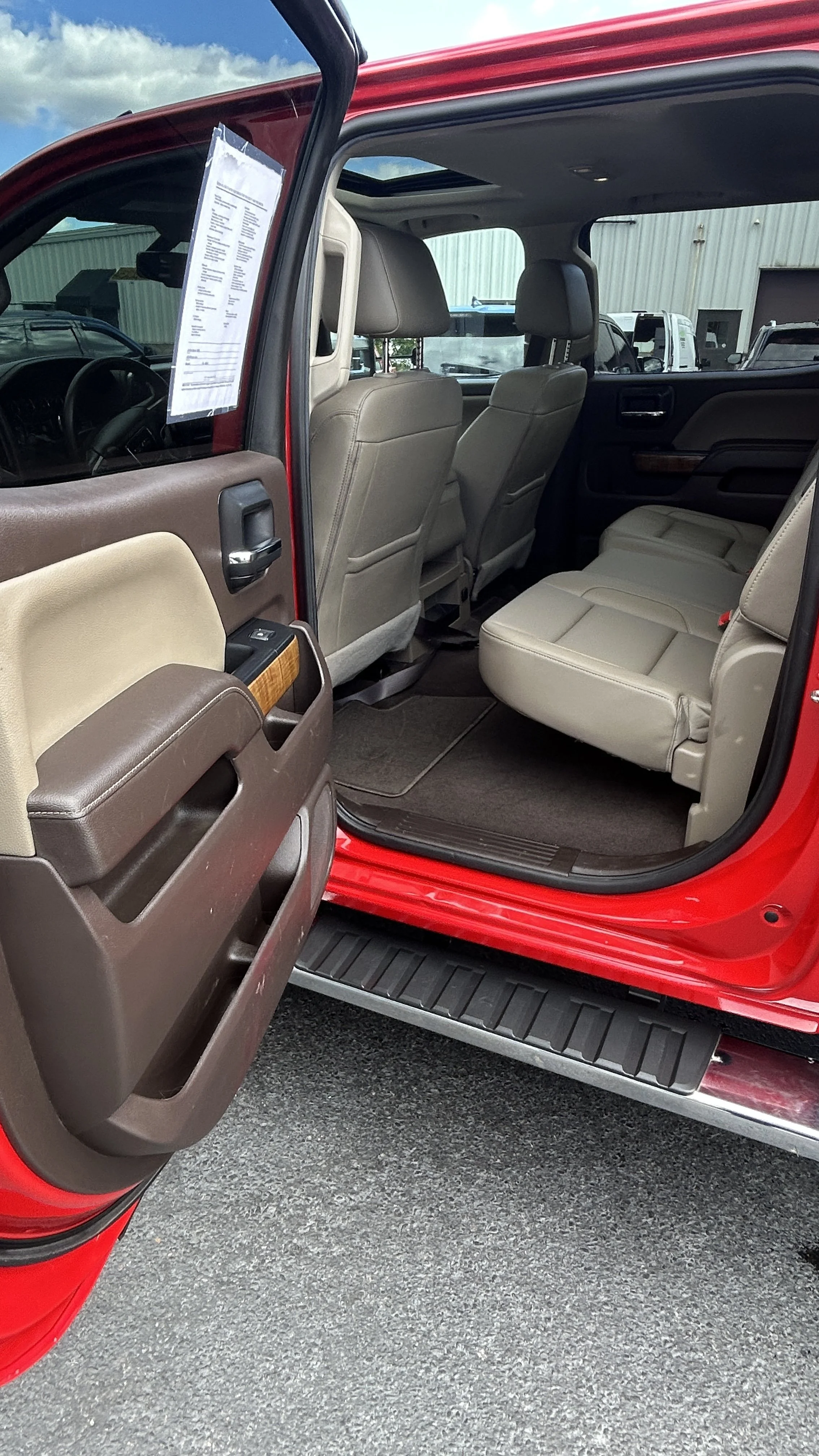 Inside of a red vehicle showing beige and brown interior seats, with the passenger-side door open revealing the car's backseat area and surrounding environment, including other vehicles and a building in the background.
