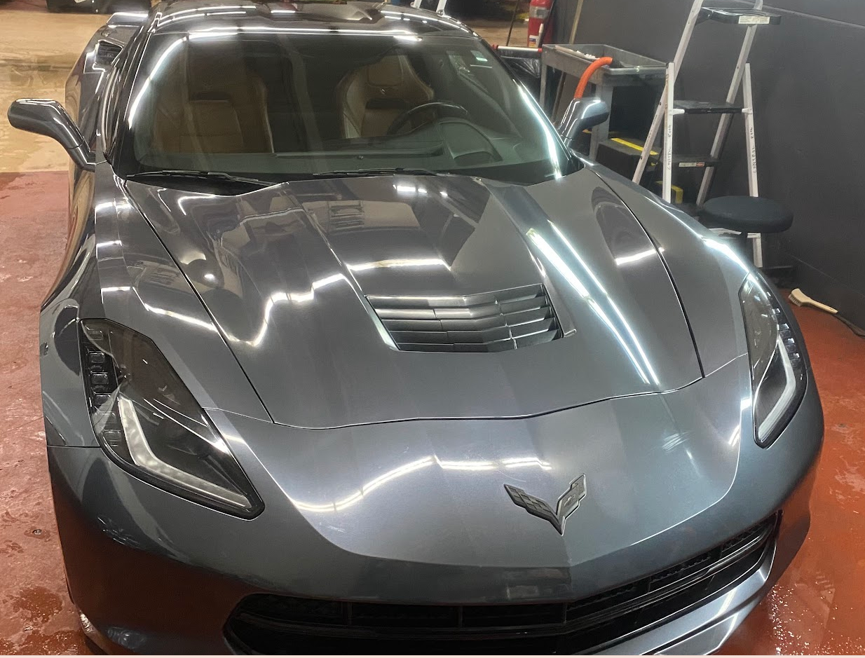 A silver Chevrolet Corvette sports car parked inside a garage, with reflections of fluorescent lights on its surface and a ladder and tools in the background.