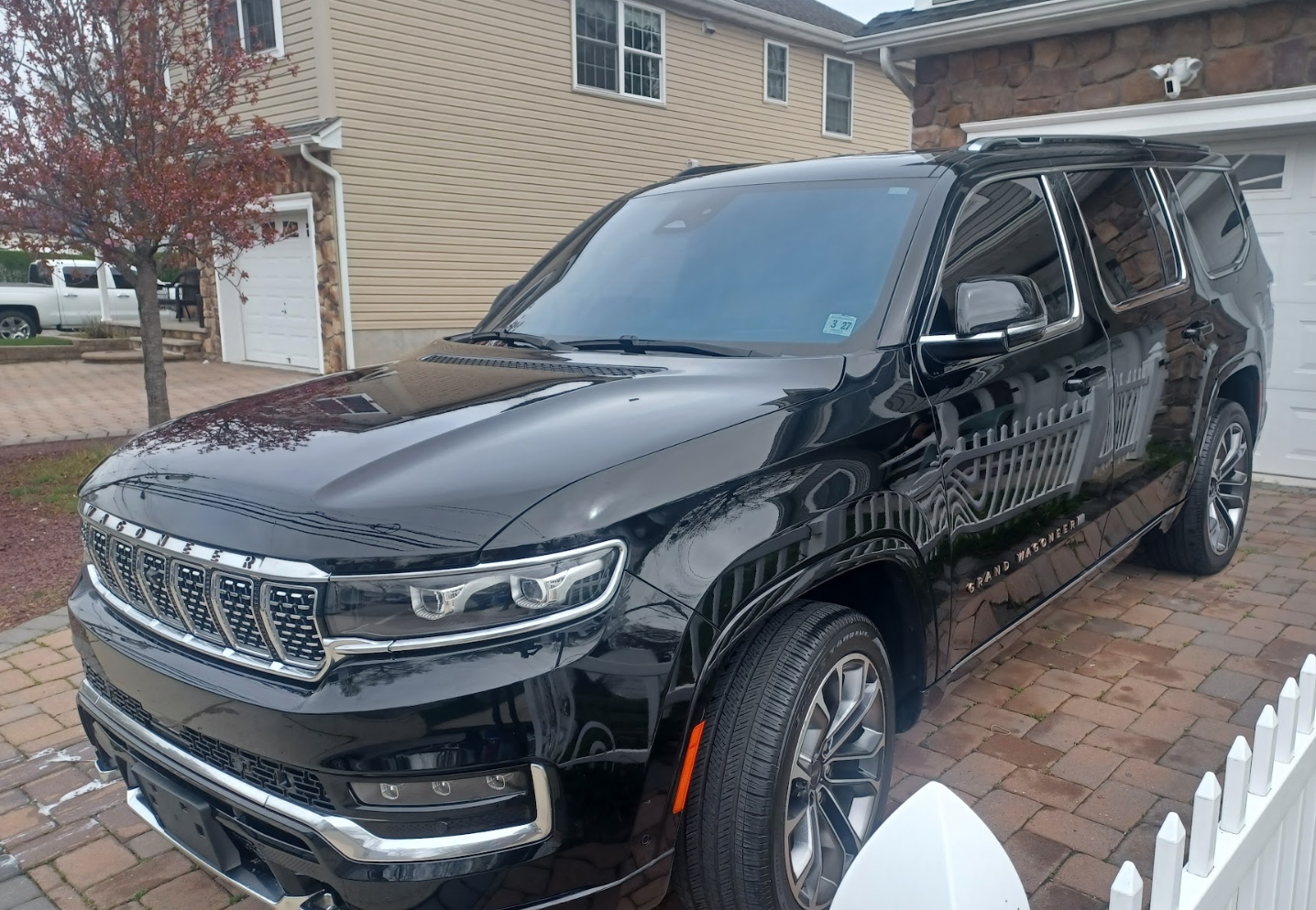 A black Lincoln Grand Crossover SUV parked on a brick driveway in front of a house with beige and stone exterior walls. There is a tree with red leaves nearby and a white picket fence in the foreground.