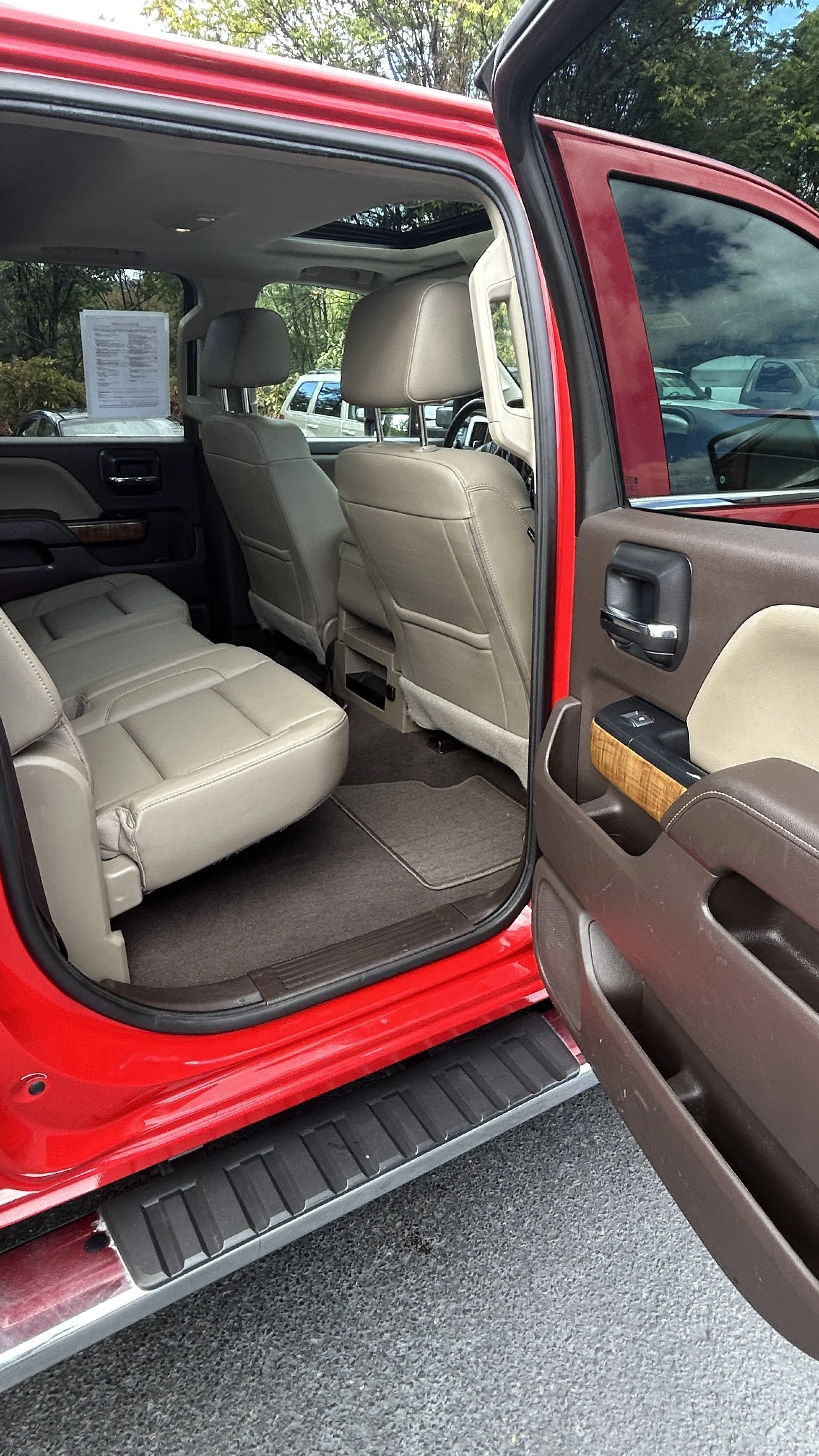 Inside a red pickup truck with tan leather seats, view from open door showing the interior including the back seat, front seats, and part of the dashboard. Outside are other parked vehicles and trees.