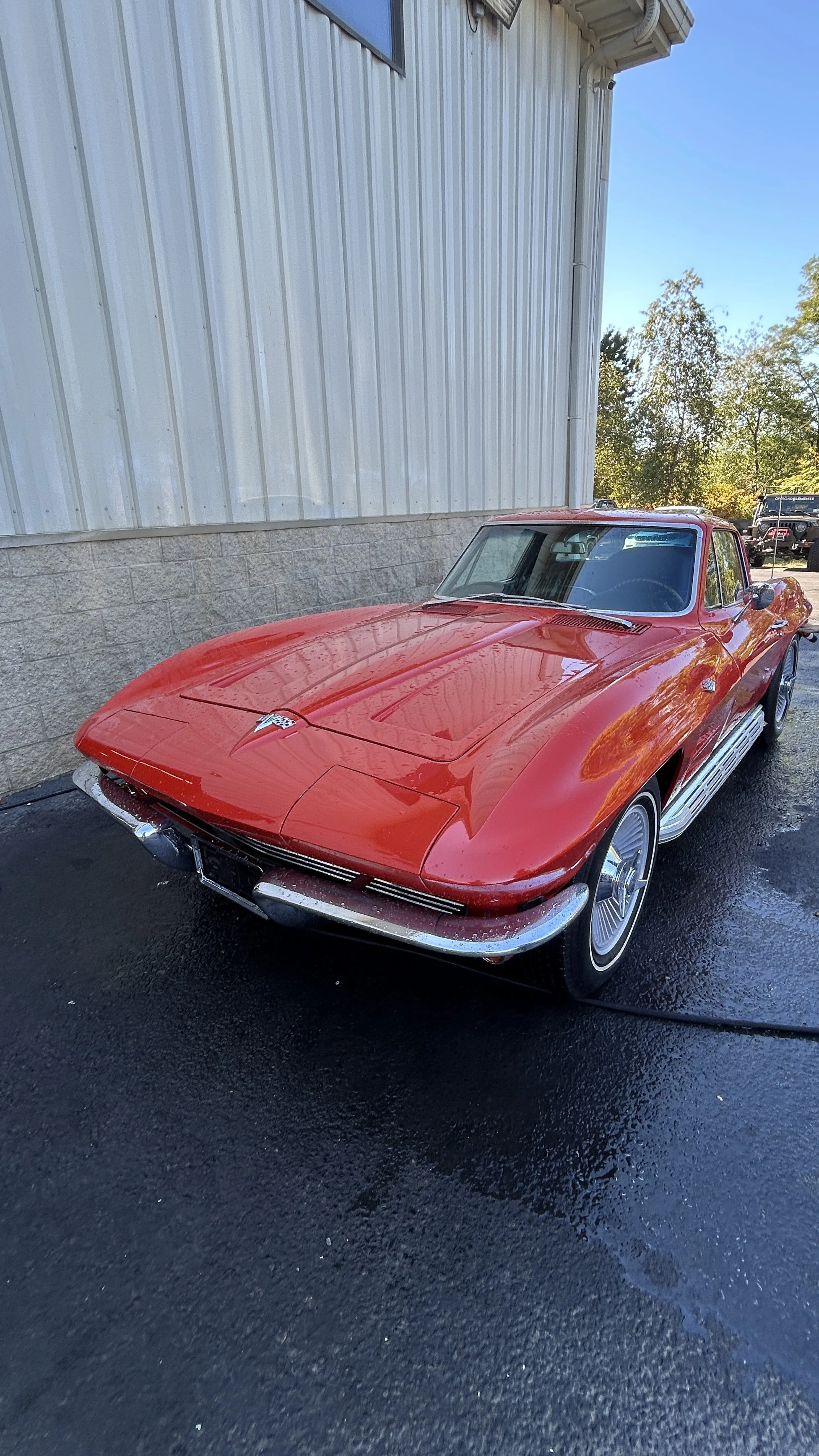Red vintage Chevrolet Corvette parked on wet asphalt outside a building with white siding.