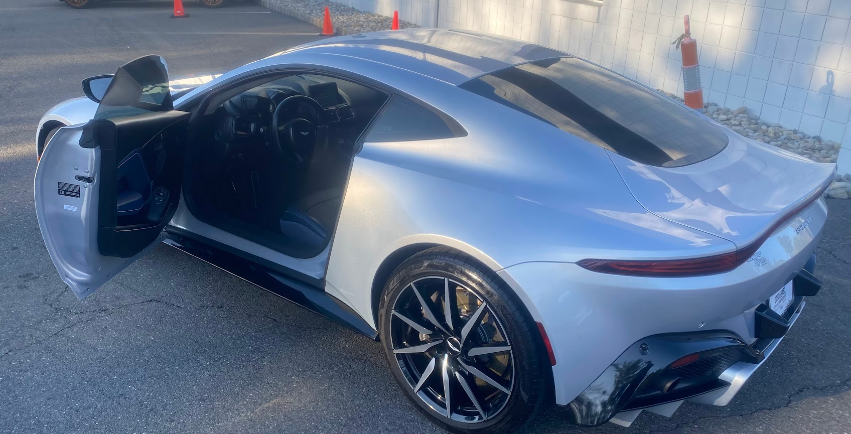 A silver sports car with its driver's side door open, showing the interior and a sleek, modern design, parked on asphalt near a white tiled wall and orange traffic cones.