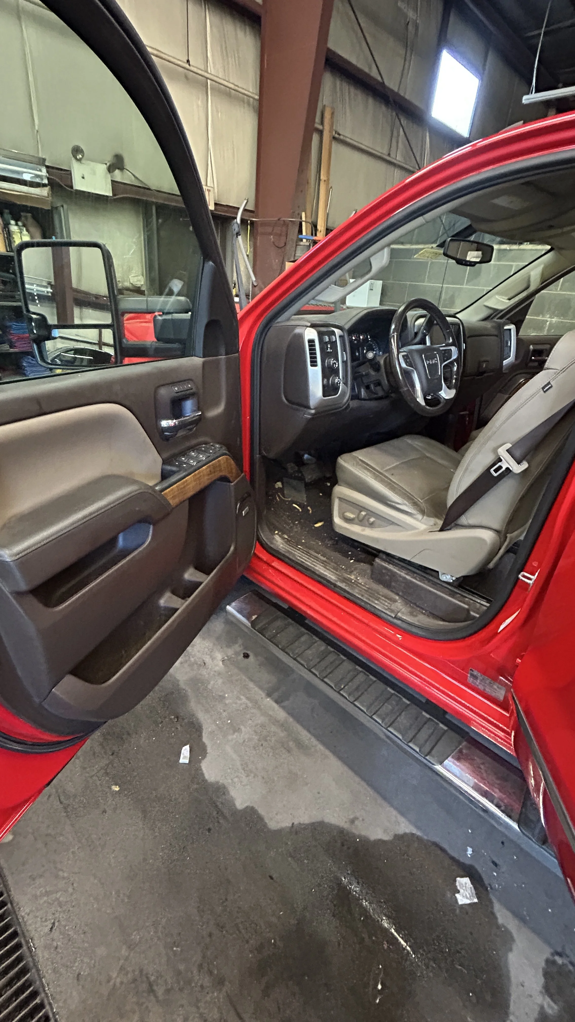 The interior of a red pickup truck showing the driver's seat, steering wheel, dashboard, and part of the door panel, with a garage-like background.