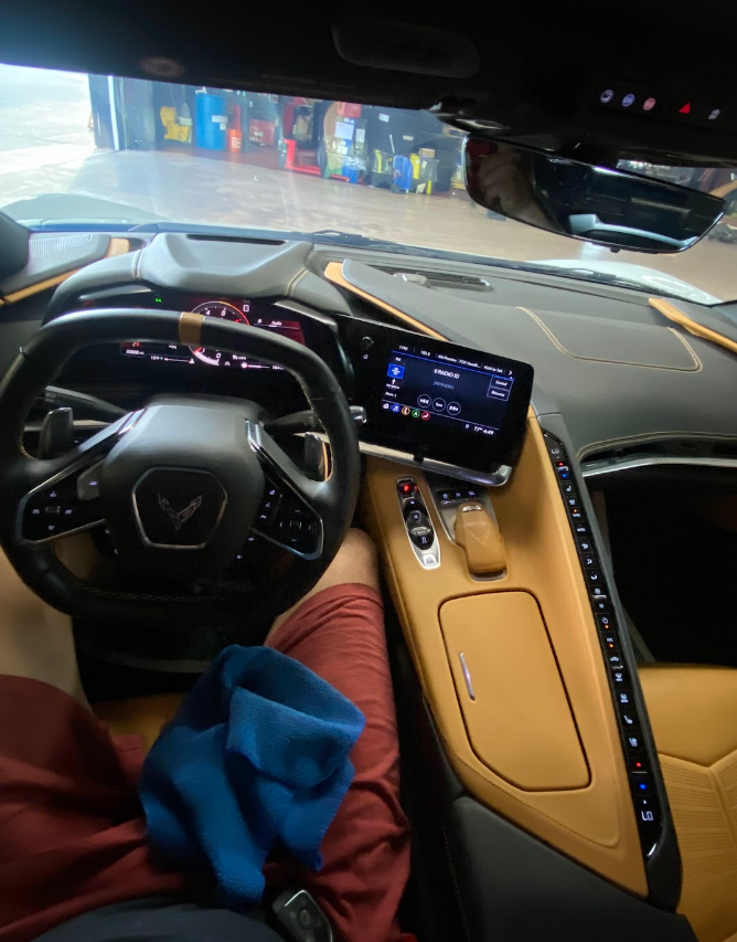 The image shows the interior of a luxury vehicle, with a patterned steering wheel, digital dashboard, large touchscreen display, and beige dashboard and seats. There is a person sitting in the driver's seat wearing red shorts and a blue cloth in thei