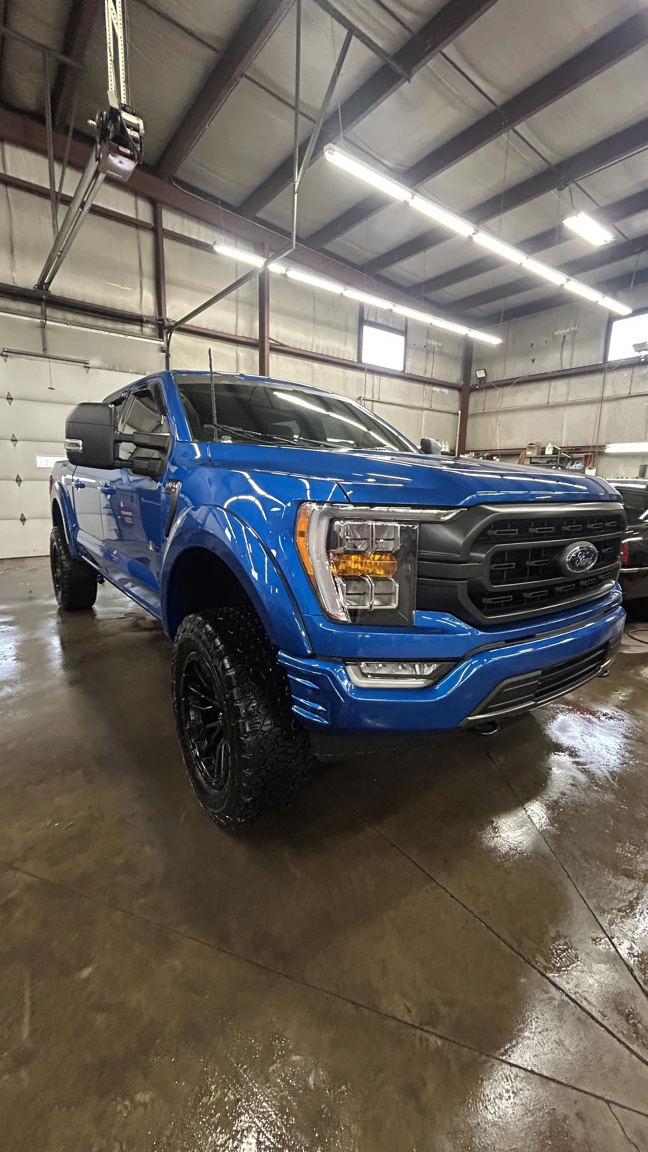 A blue Ford pickup truck parked inside a large industrial garage with high ceilings and fluorescent lighting.