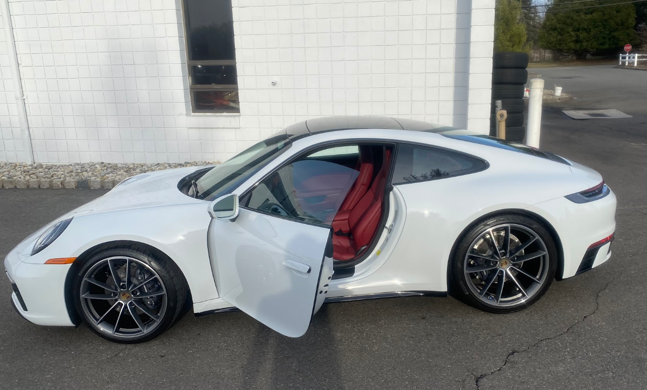 A white Porsche sports car with its driver's side door open, revealing red interior seats. The car is parked outdoor on asphalt near a white building with a window and a stack of tires.