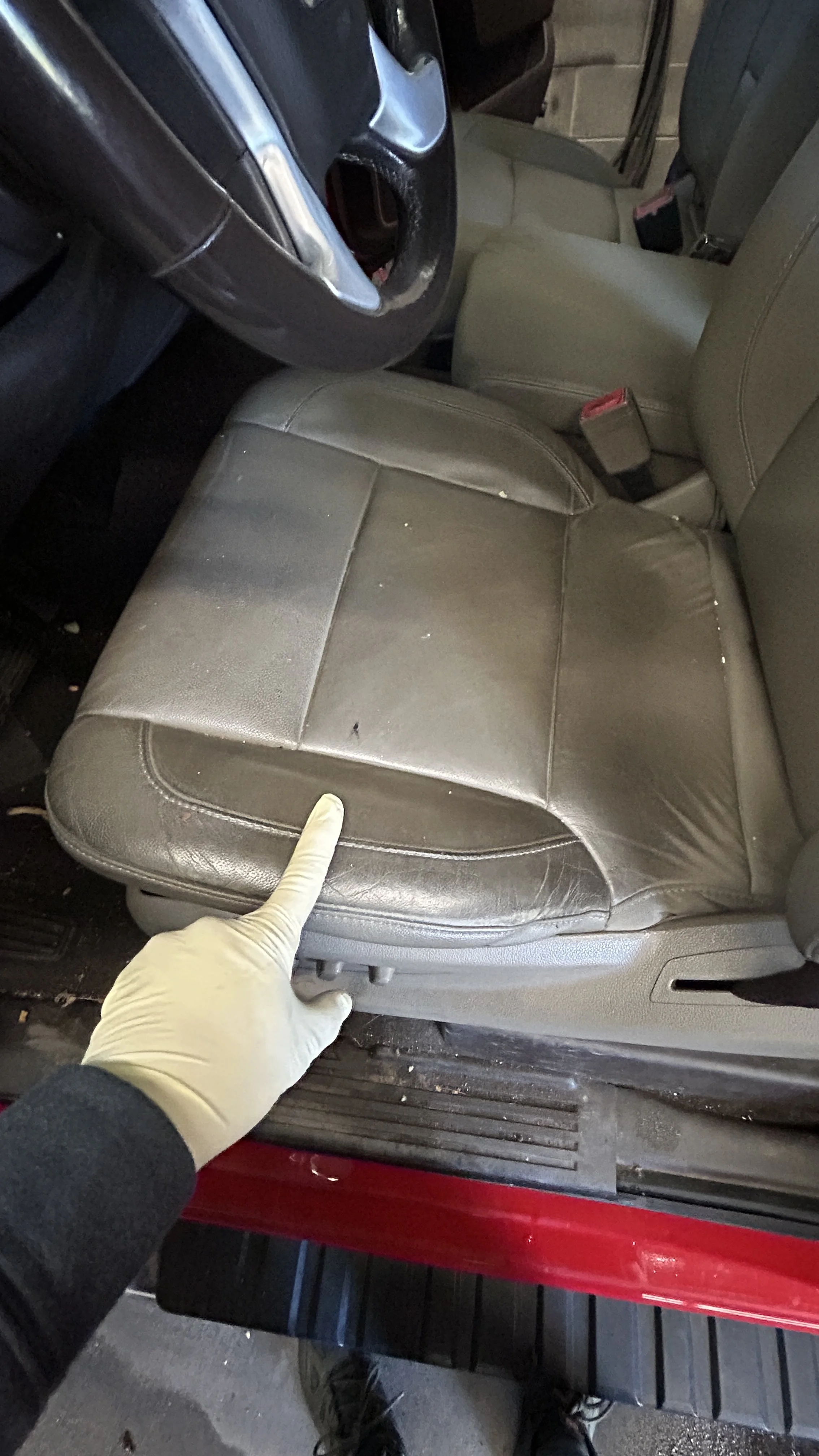A person wearing a white glove pointing at a worn, cracked spot on a car's gray leather passenger seat.