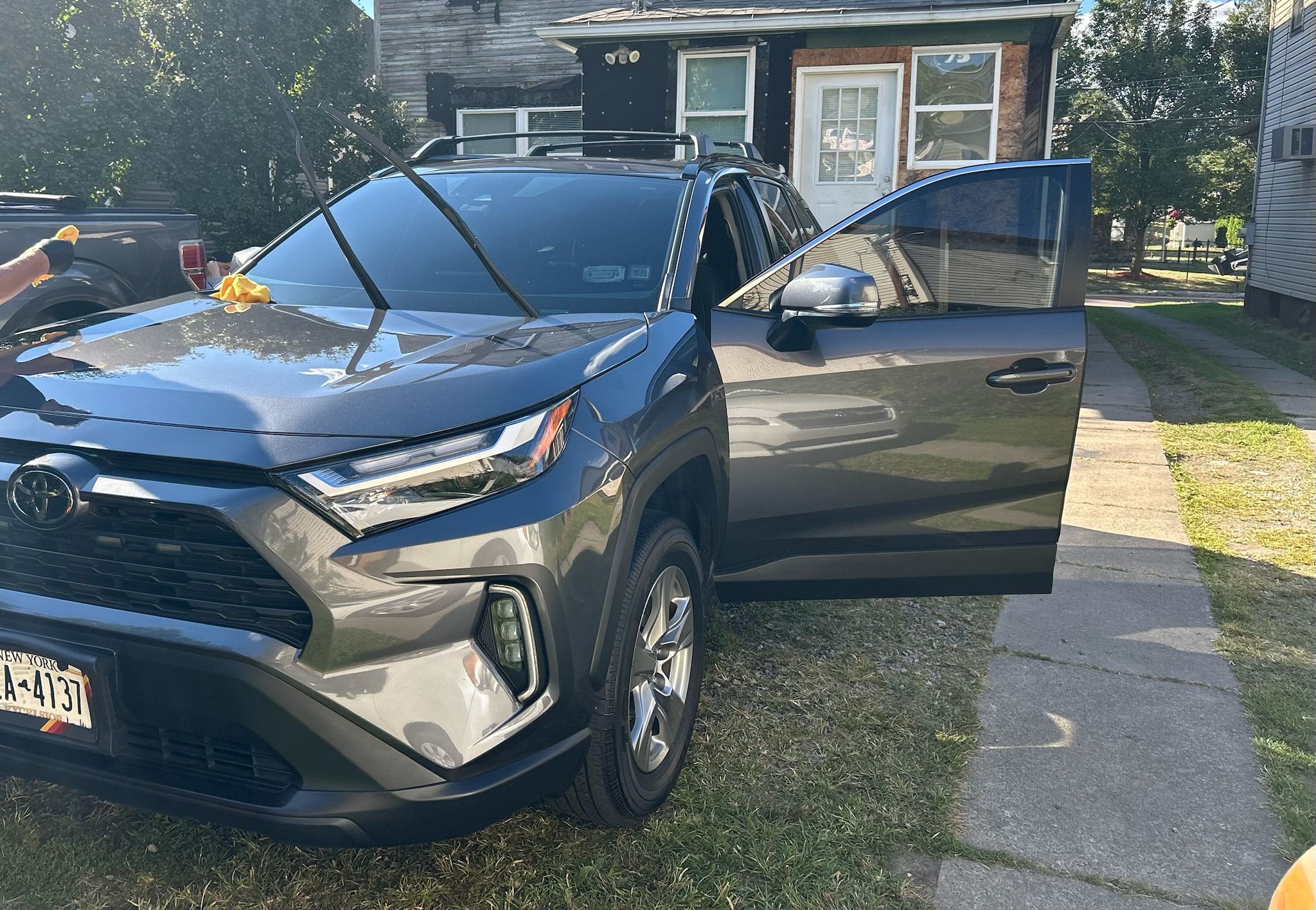 A gray Toyota RAV4 parked on a grassy patch beside a sidewalk, with its front facing left and the driver's side door open. There's a person in the background on the left wearing gloves and handling a window squeegee or cloth. The background includes a wooden house with a white door and a window, and other houses and trees are visible.