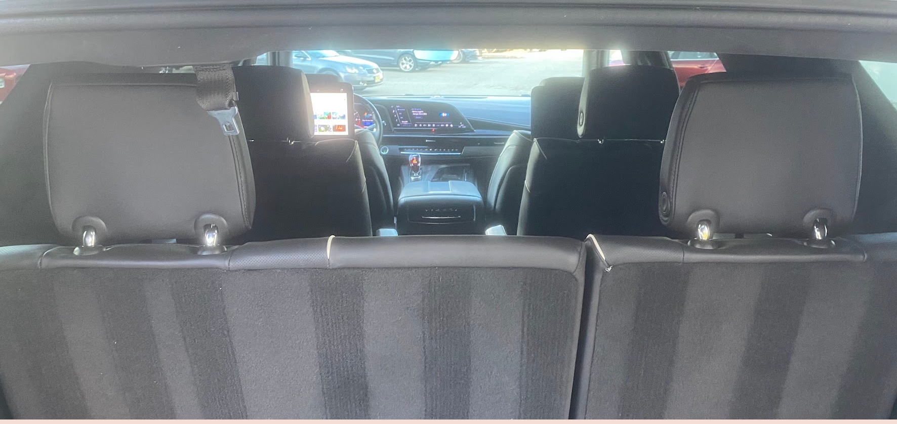 View from the back of a car showing the interior with black seats, headrests, a touchscreen display on the dashboard, and a parking lot outside with other vehicles.