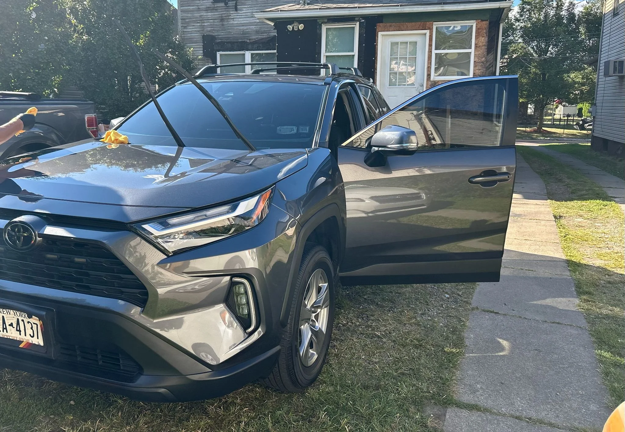 A gray Toyota vehicle parked on a patch of grass and concrete in front of a house, with the front passenger door open. Someone is cleaning the car's windshield with a yellow cloth, and a hand wearing a black glove is visible holding the cloth. There 