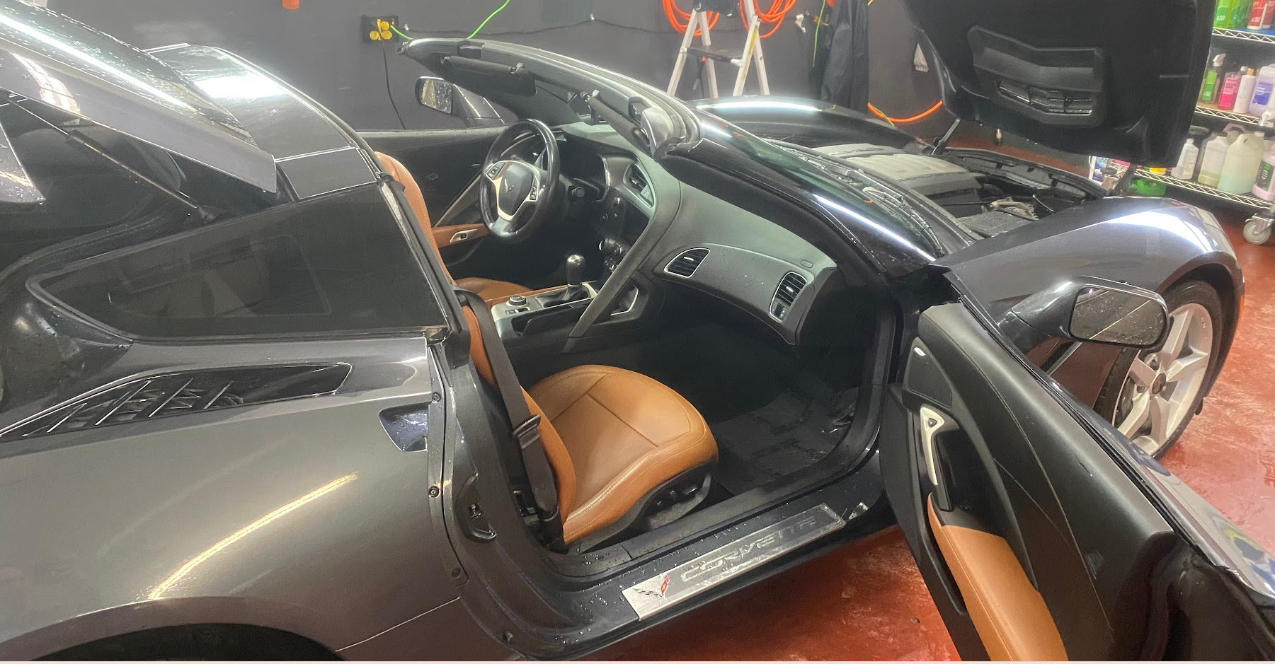 A gray convertible sports car with tan leather seats parked inside a garage. The car's hood is open, exposing the engine, and the driver's side door is open, revealing the interior. Some garage shelves with bottles and cleaning supplies are visible i