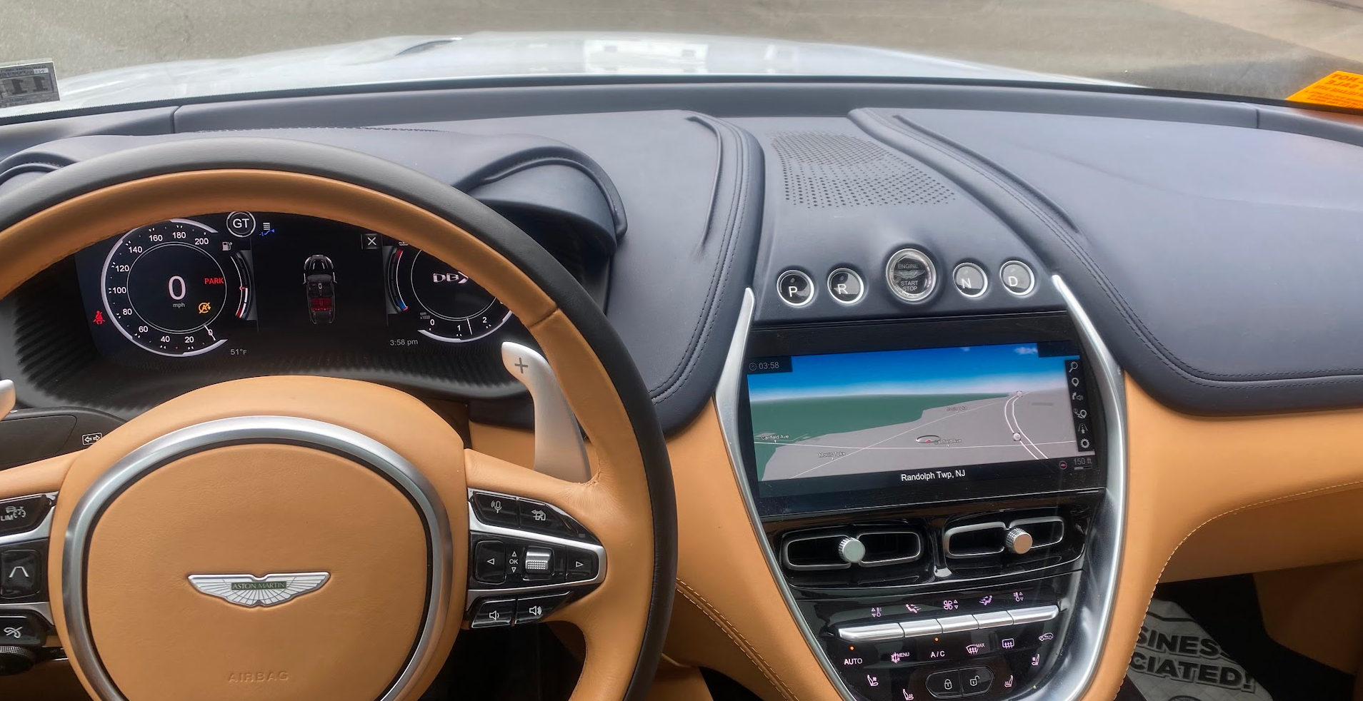 Interior view of an Aston Martin car dashboard showing a digital speedometer, navigation screen displaying a map of Randolph Twp, NJ, and various control buttons and dials.