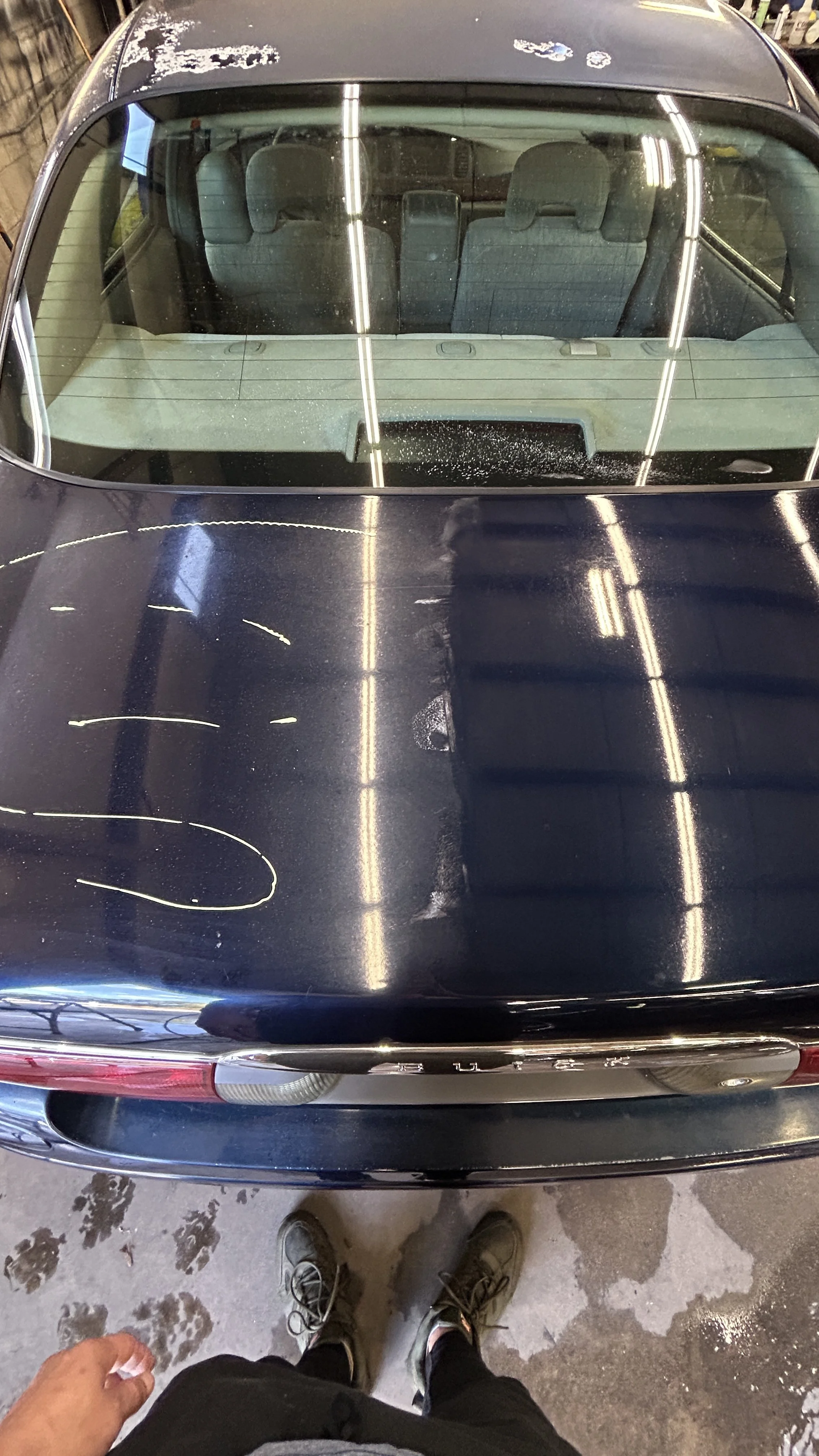 Rear view of a black car with a shiny, slightly dusty hood, viewed from above. The photo includes the photographer's feet in sneakers and a hand holding a tissue or cloth. The ground shows some wet patches and tire tracks.