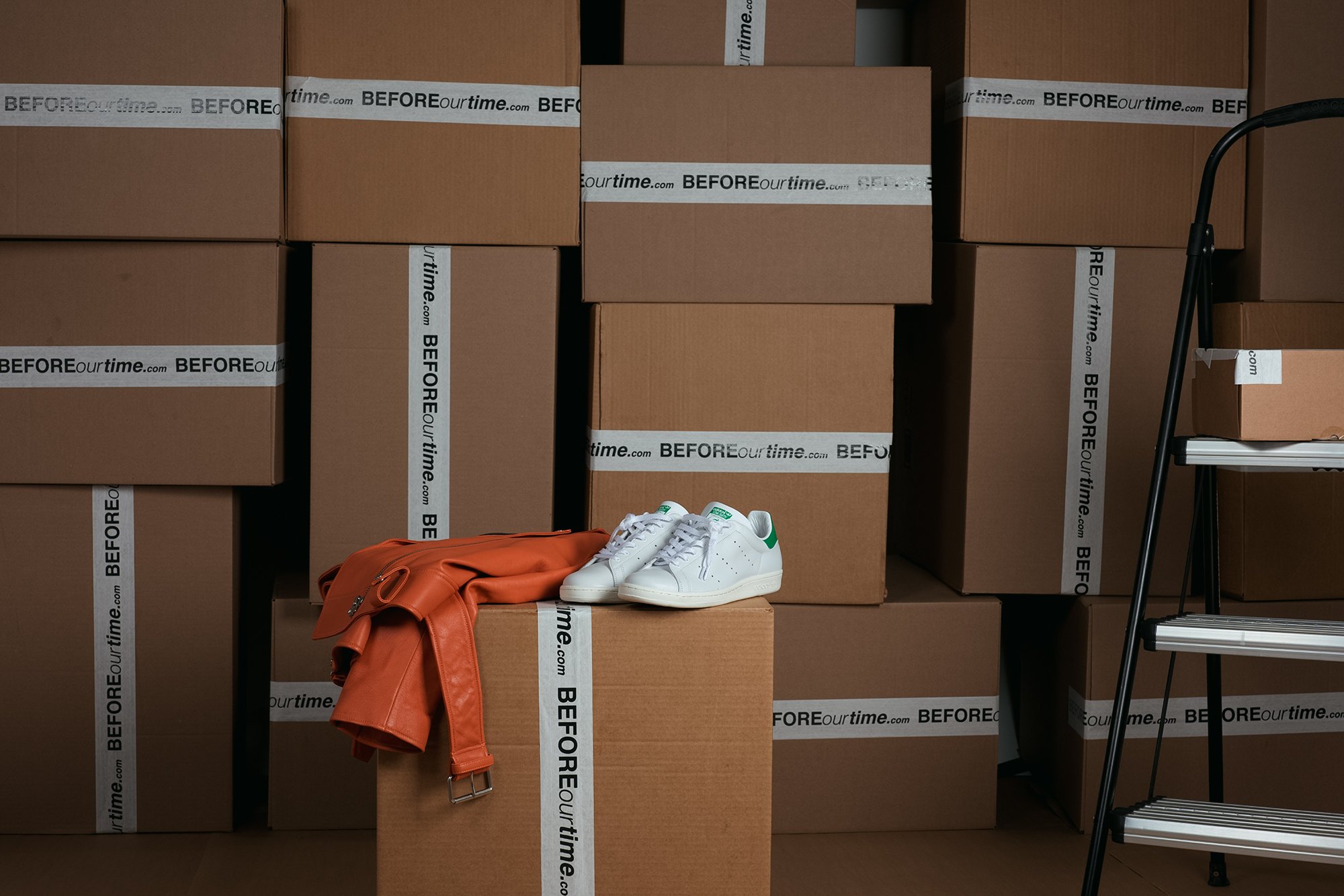 Stack of cardboard boxes with 'BEFORE ourtime.com' shipping labels, a pair of white sneakers, and an orange jacket draped over a box, with a metal stepladder on the side.