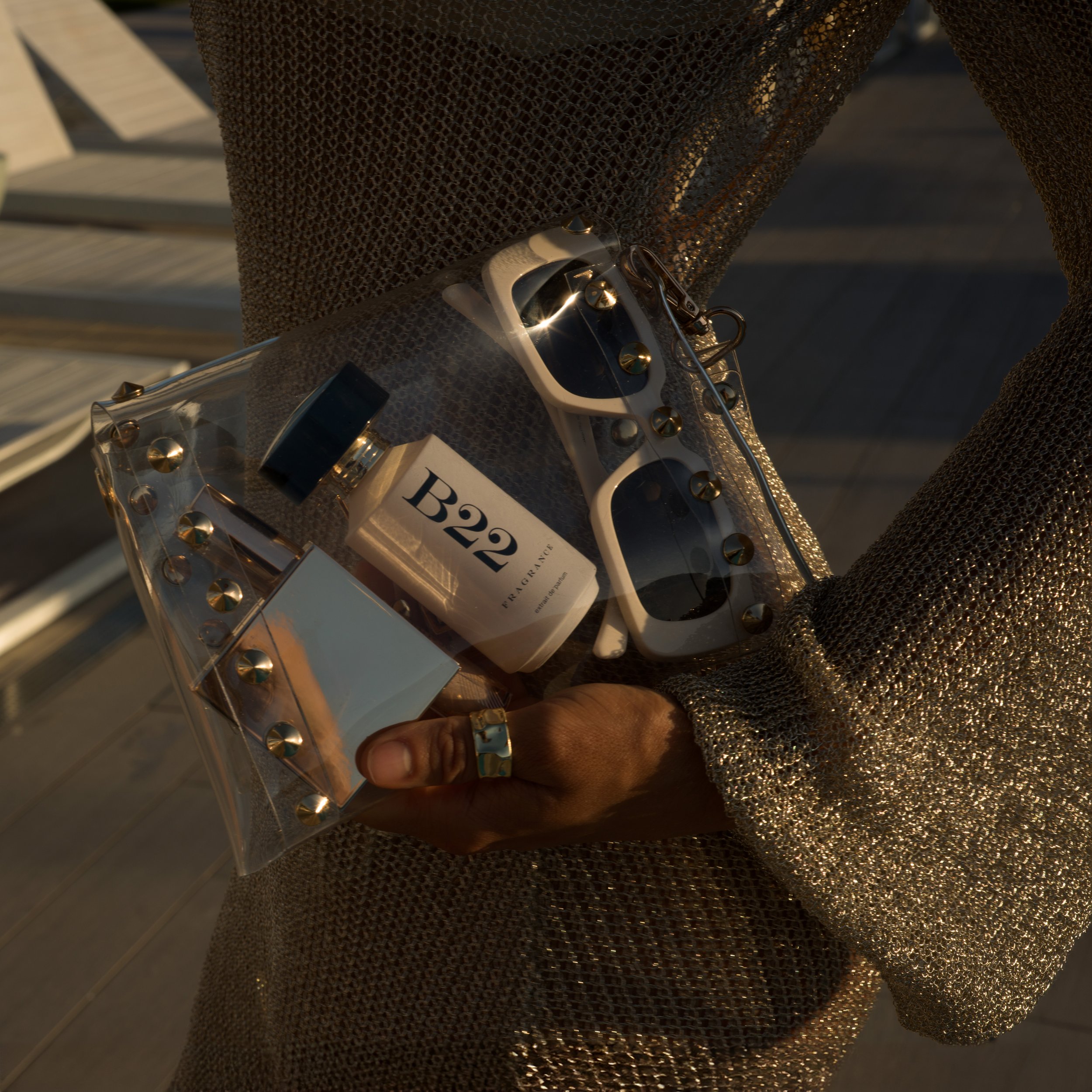 A person holding a clear plastic bag with gold studs, a bottle of B22 perfume, a pair of white sunglasses, and another small silver container. The person is wearing a metallic, gold, long-sleeved top and a gold ring.