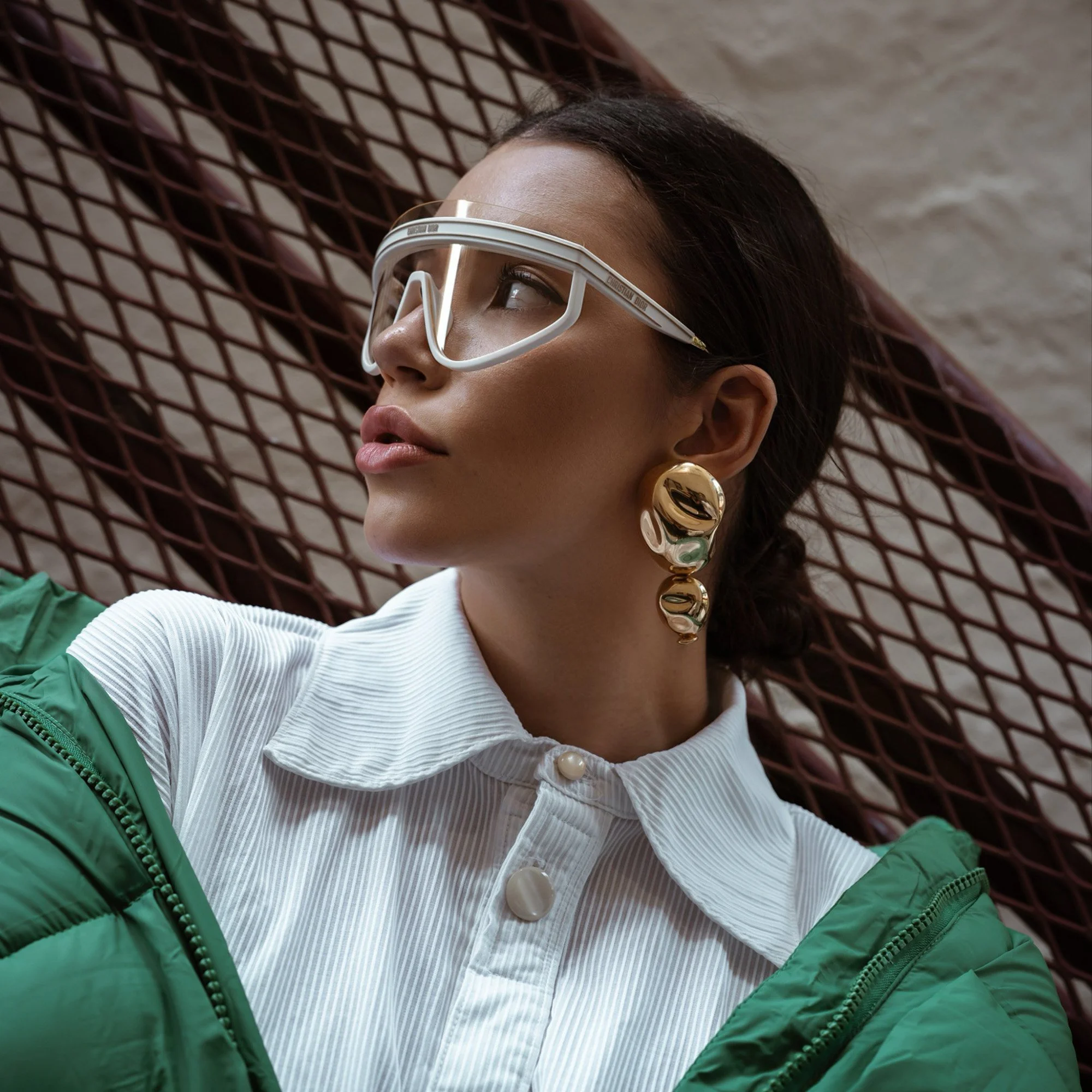 A woman wearing large gold statement earrings, white button-up shirt, green jacket, and white goggles, posed against a brick wall background.