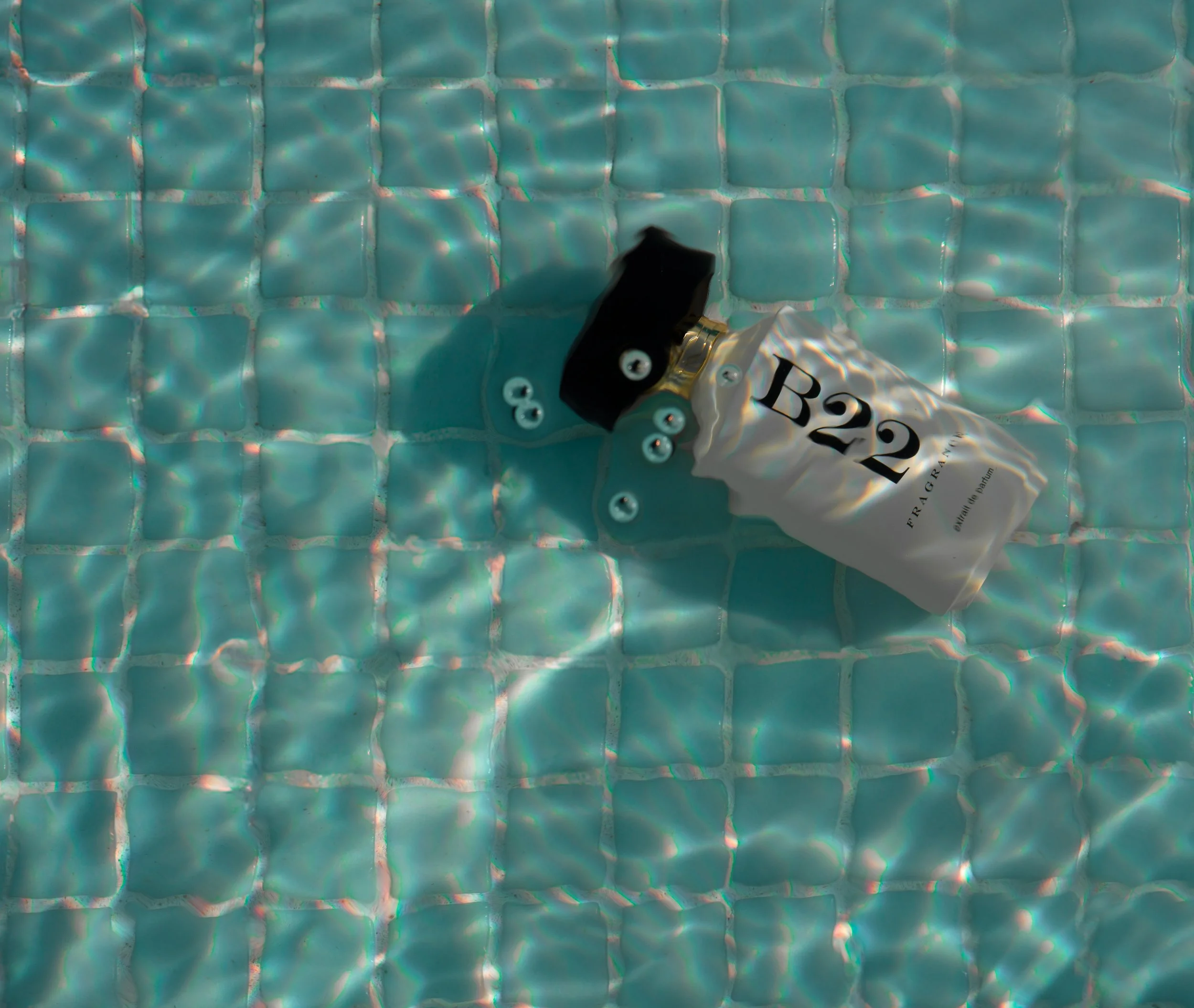 A partially squeezed white bottle with black text that reads 'B22'. The bottle is surrounded by small white bubbles and is lying on a tiled surface with a turquoise, mosaic-like pattern. The lighting creates bright reflections on the tiles and on the