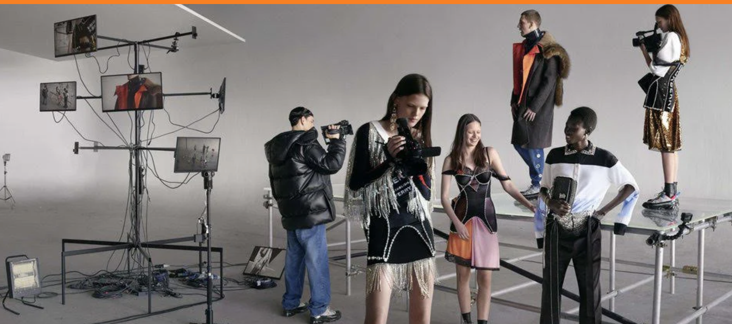 Fashion models in colorful outfits posing on a raised platform during a photoshoot, with photographers and studio equipment around them.
