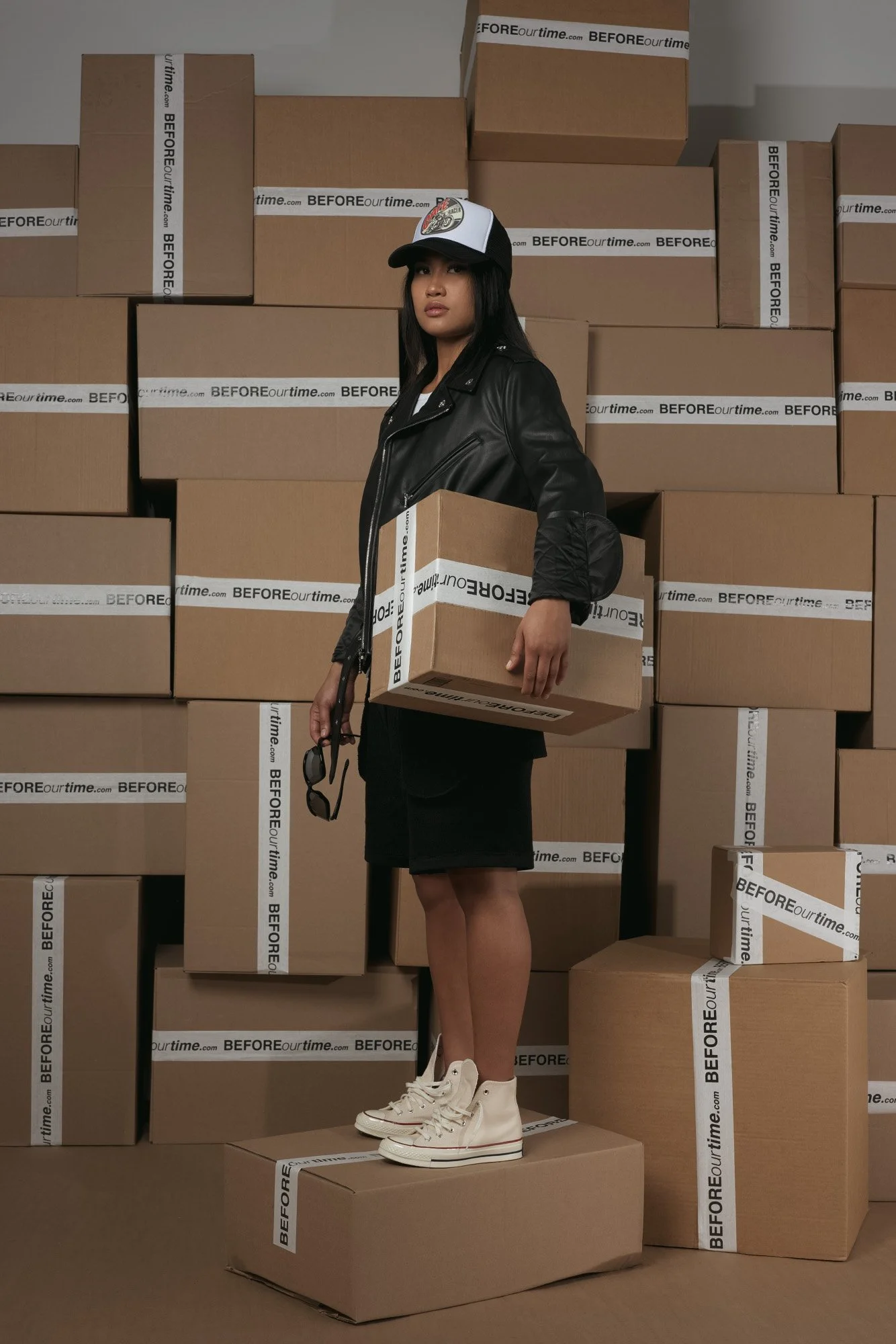 A young woman with dark hair in a black leather jacket and white high-top sneakers standing among stacked brown boxes. She holds a box and a pair of sunglasses, and wears a black and white cap. The background and boxes are branded with "BEFOREourtime