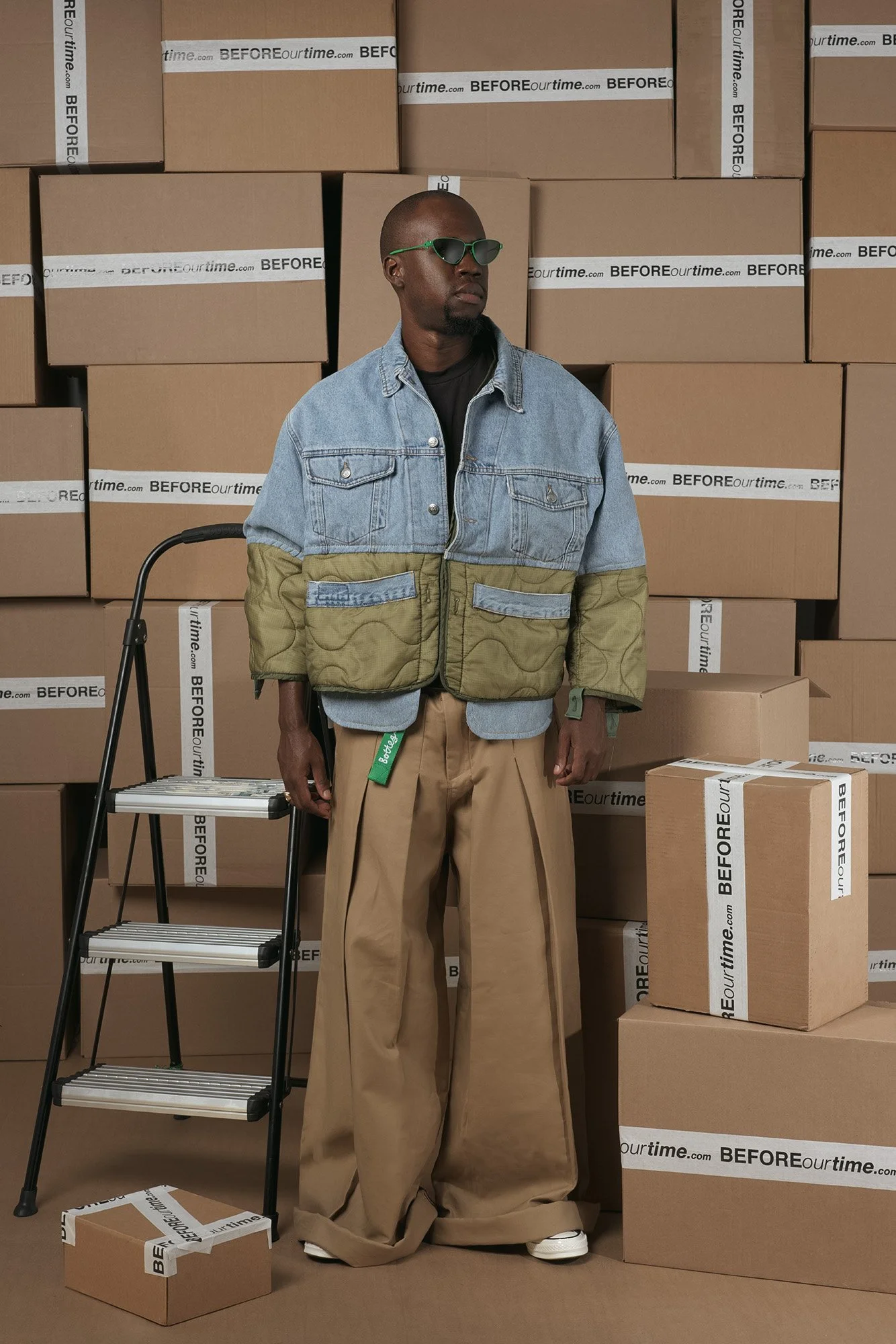A man wearing sunglasses, a black shirt, a color-blocked denim and khaki jacket, and beige wide-leg pants stands among cardboard boxes and a small black folding ladder.