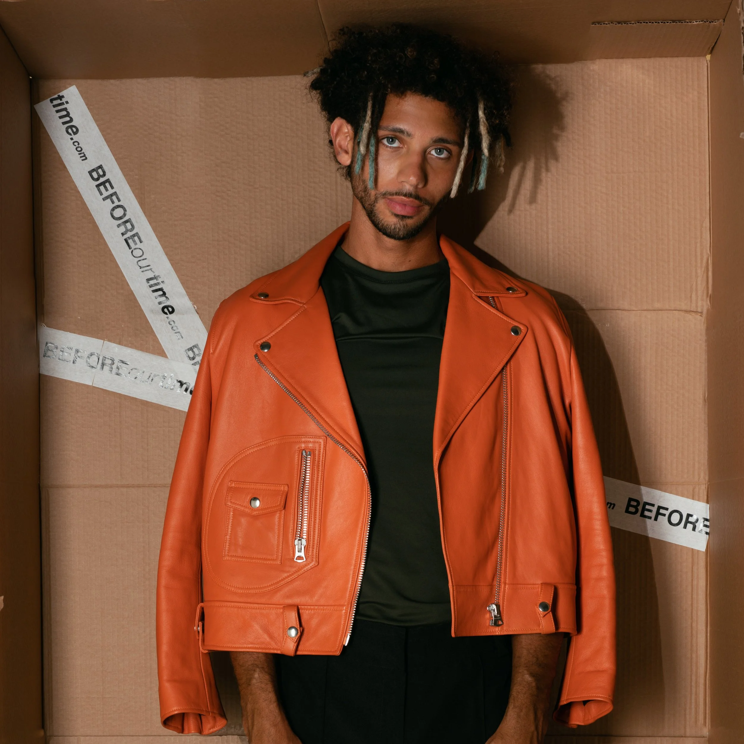 A young man with curly hair and highlights, wearing an orange leather jacket over a black shirt, standing inside a large cardboard box with 'BEFORE' tape.