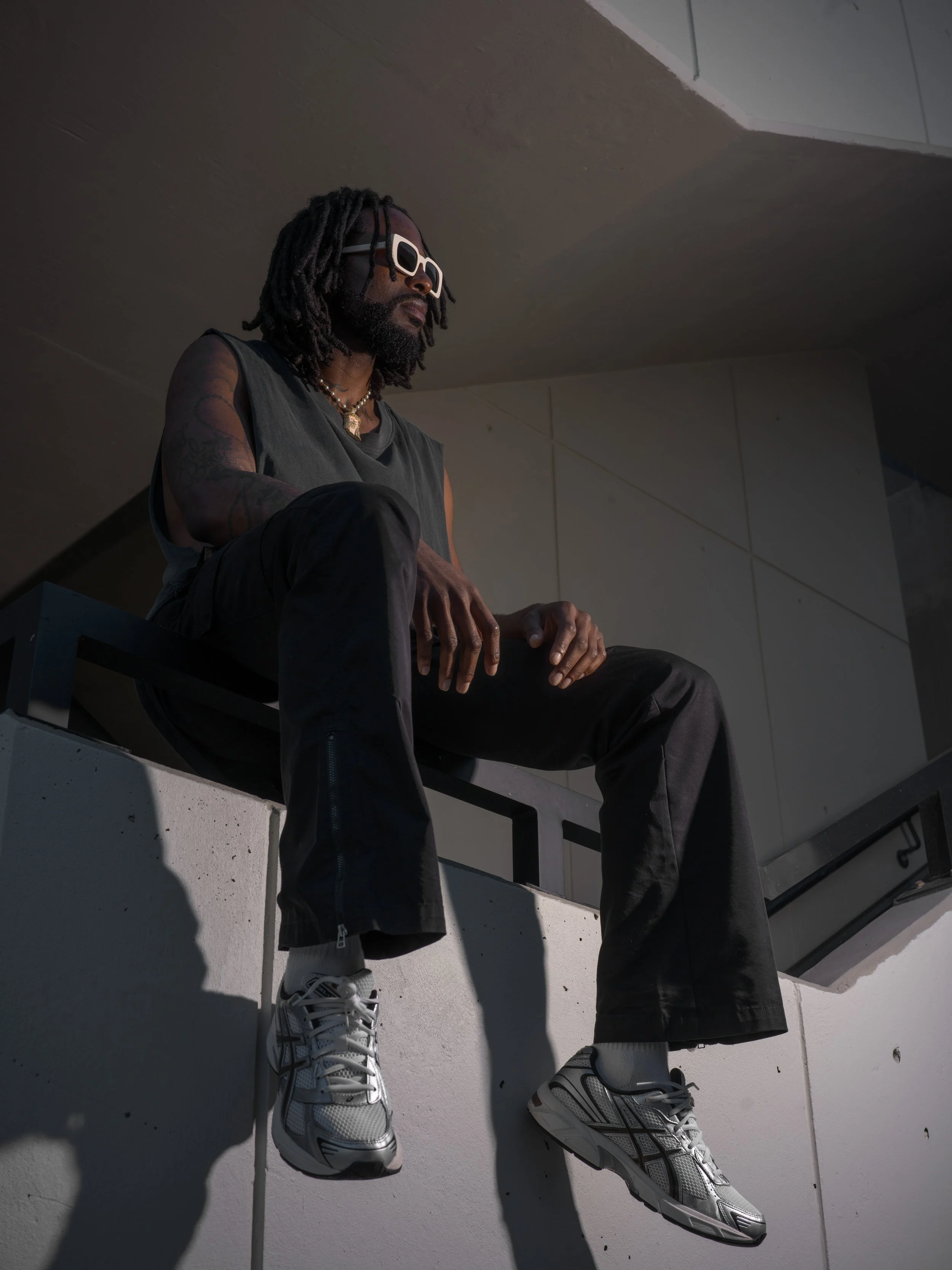 A man with dreadlocks, wearing white sunglasses, a sleeveless dark shirt, black pants, and white sneakers, sitting on a ledge in an indoor setting with shadows cast behind him.