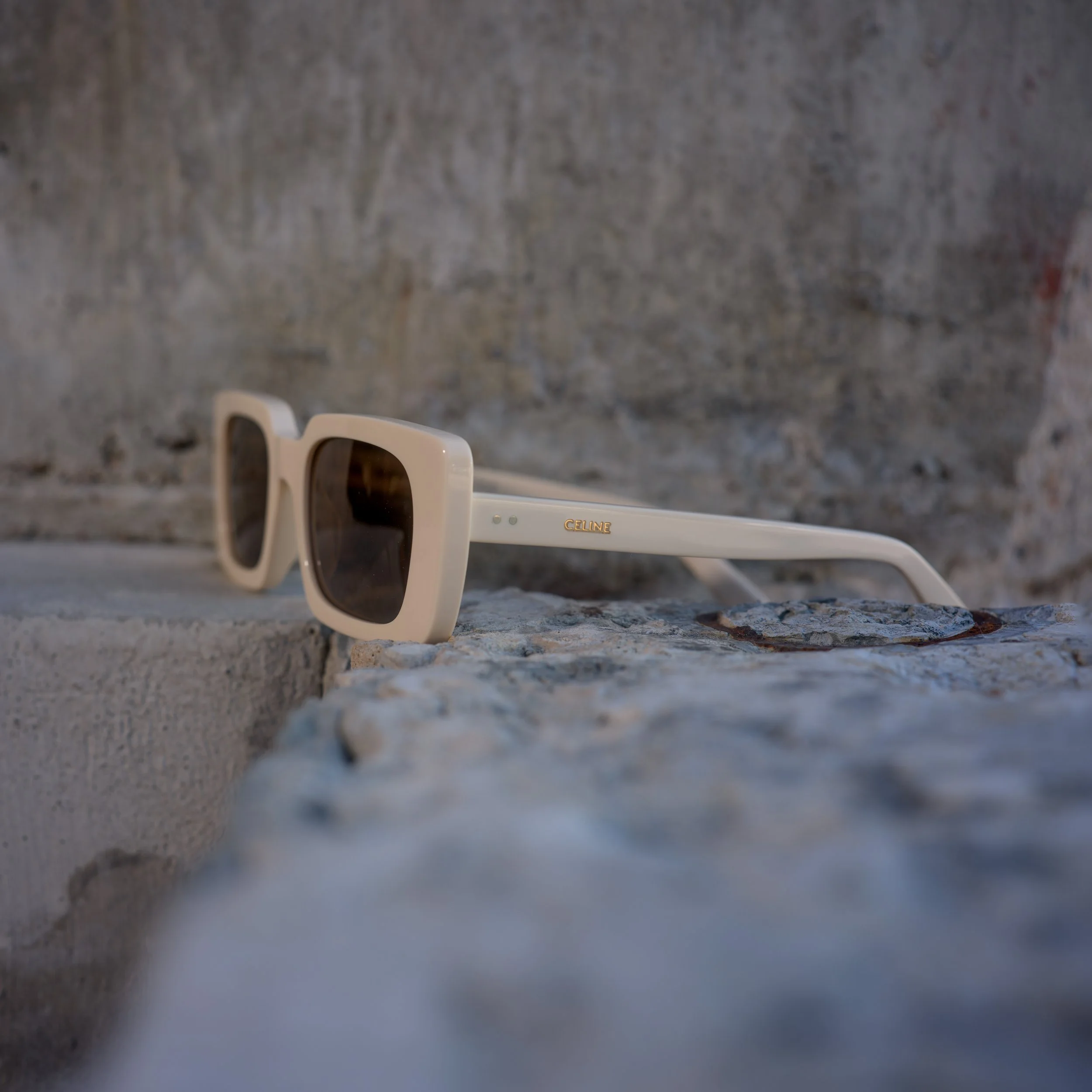 White Celine sunglasses with dark lenses resting on a rough stone surface against a stone wall background.