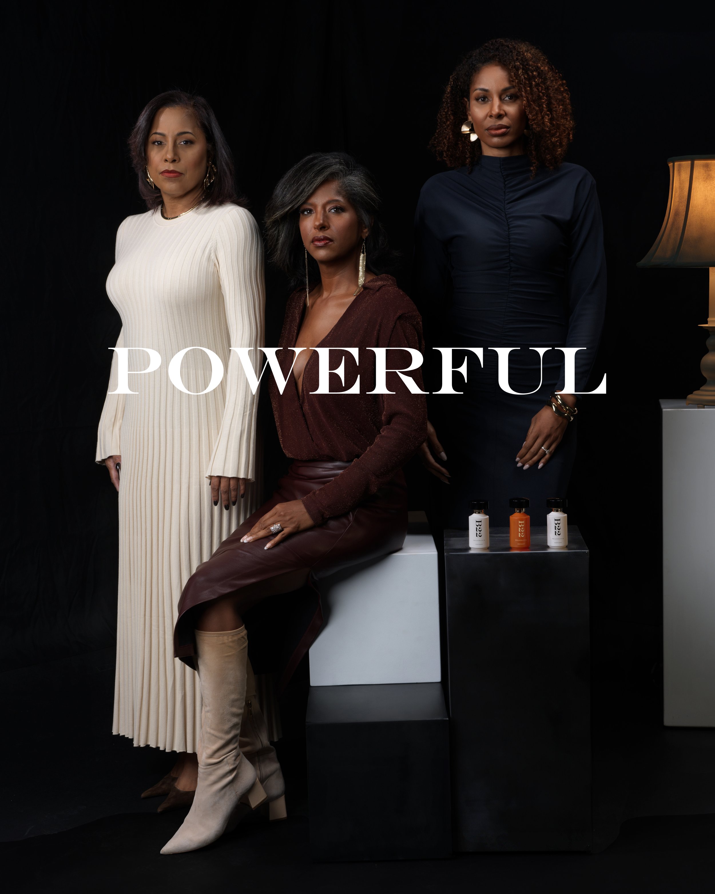 Three elegant women standing against a black background, one sitting on a black and white block, with the word 'POWERFUL' written across the image. Items such as bottles are placed on the block.