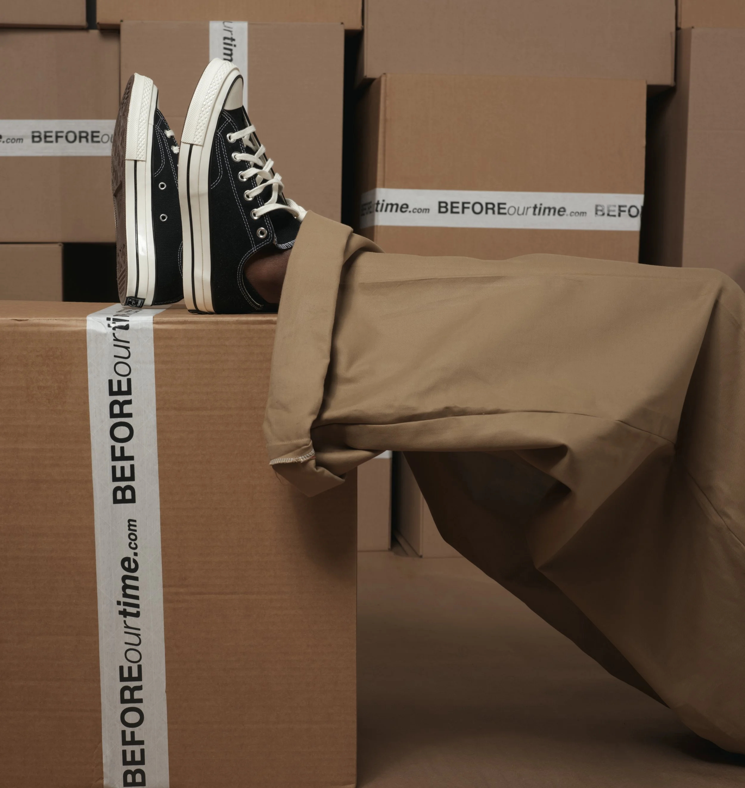 Person's legs with brown pants and tan tape, resting on a cardboard box with other boxes in the background. The boxes have 'BEFOREourtime.com' printed on the tape.