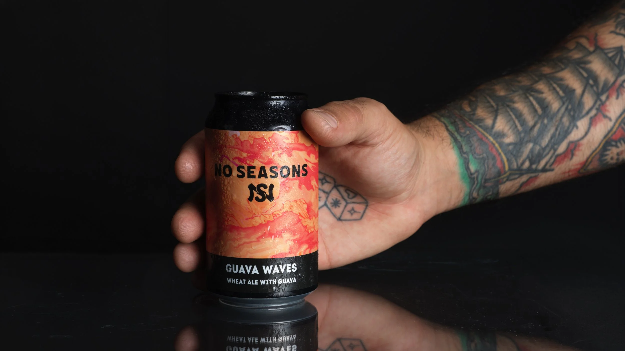 A tattooed hand holding a can of No Seasons AS, Guava Waves wheat ale with guava, against a black background.