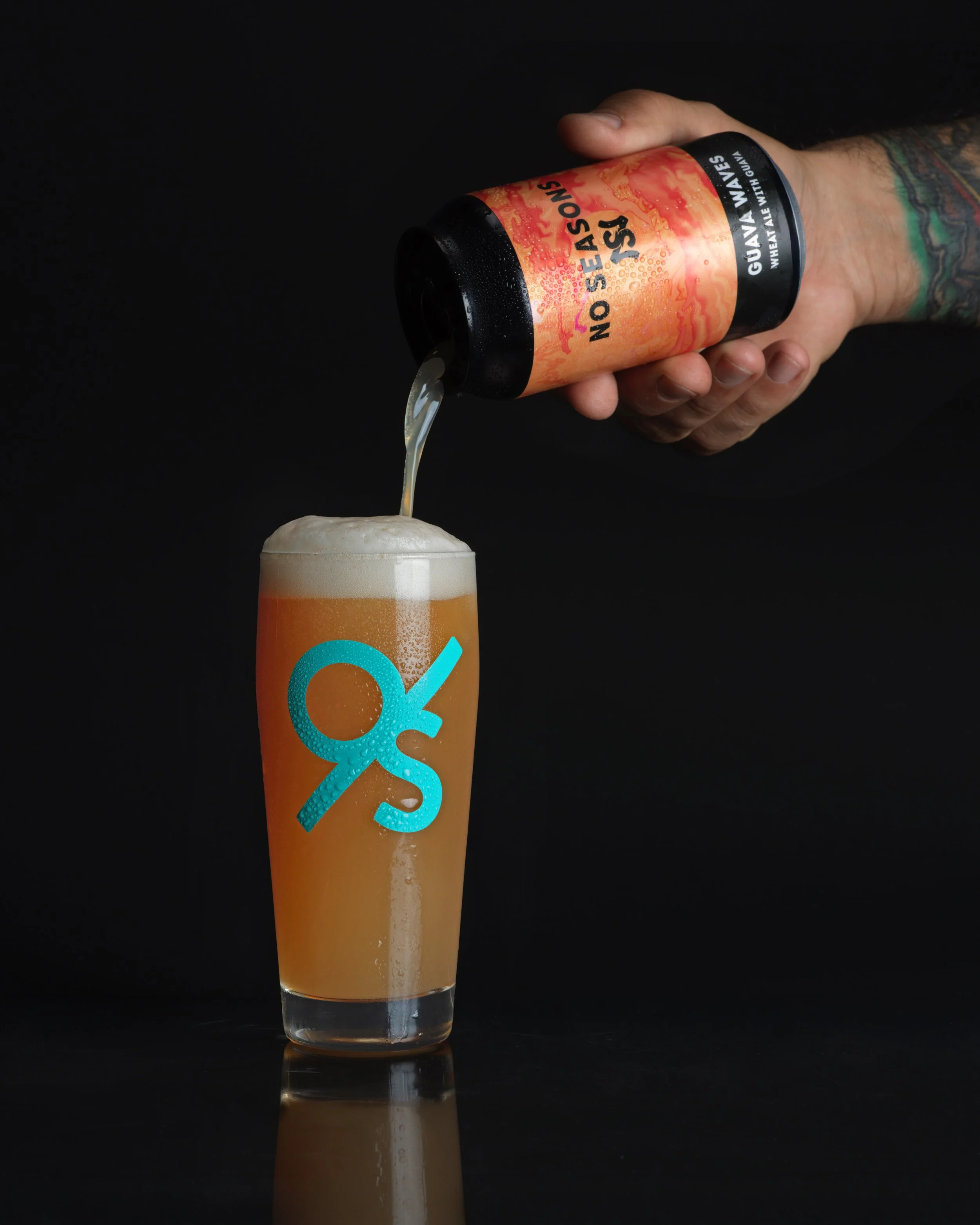 A person with tattoos pouring guava sour beer from a can into a glass with a blue logo, creating a foamy head against a black background.