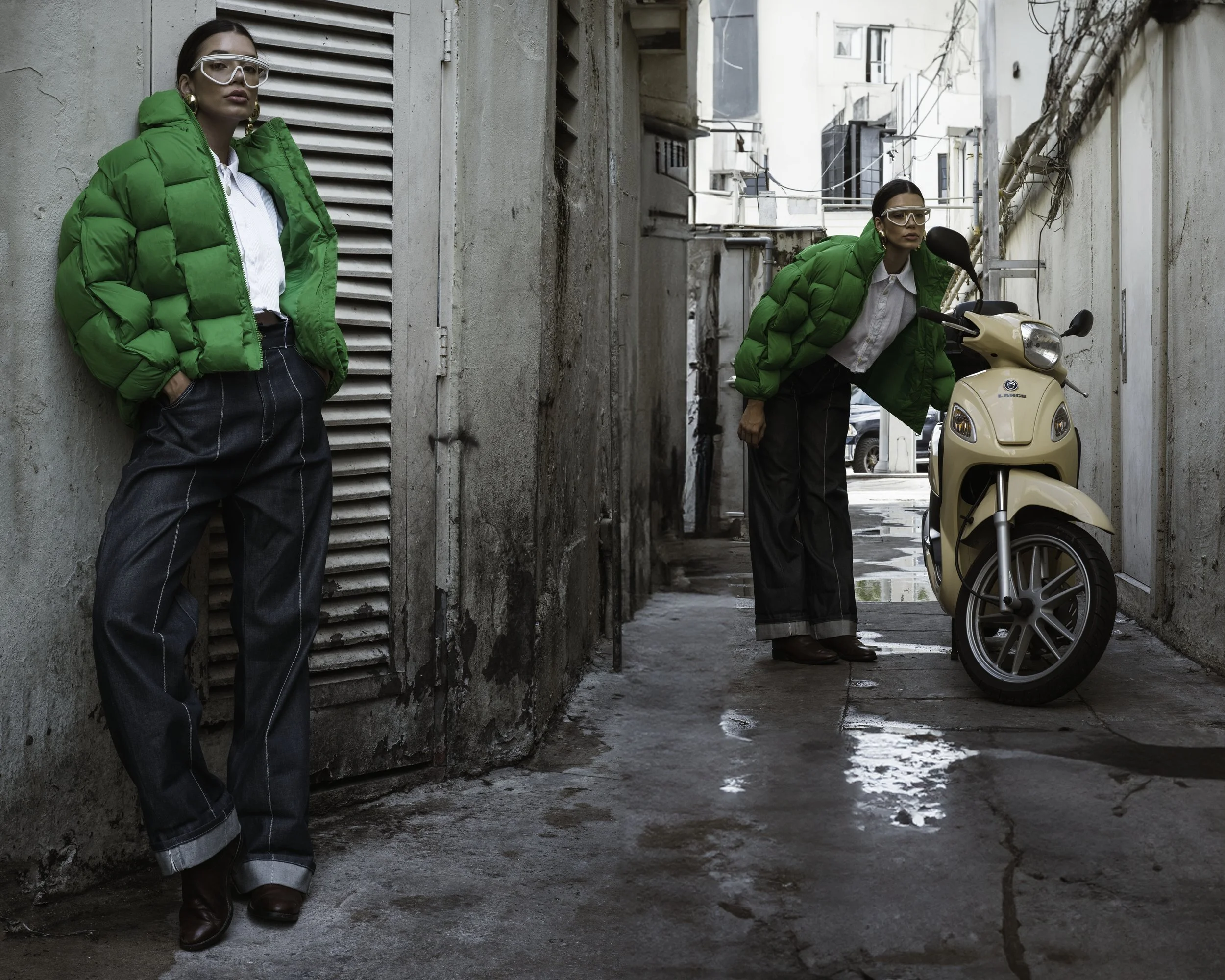 Two women in green puffer jackets and black wide-leg jeans, one standing against a wall and the other leaning over a parked beige scooter in a narrow urban alleyway.