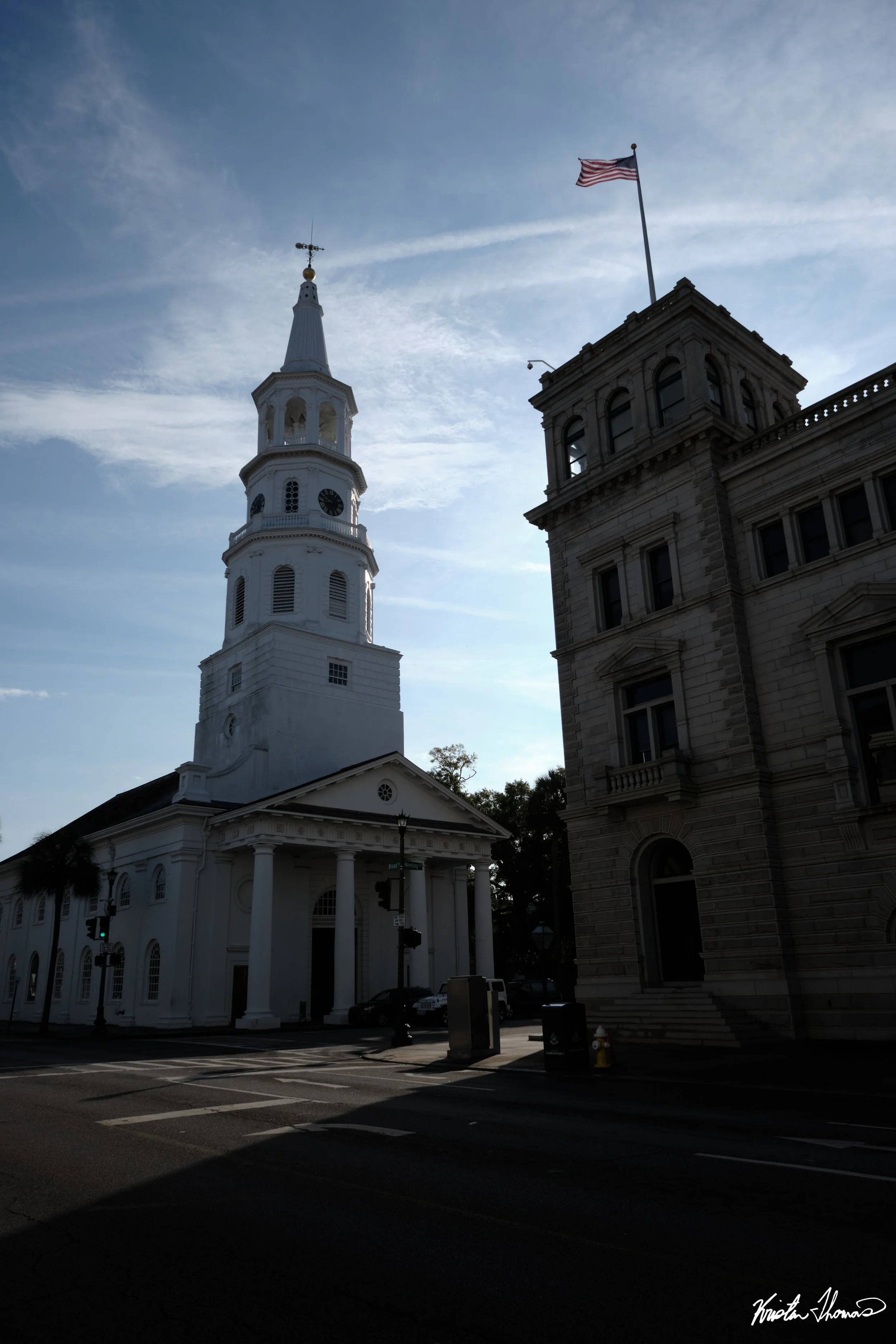 The image shows a tall white church with a spire and a clock, alongside a nearby building with a flagpole and an American flag. The sky is partly cloudy with the sun behind the buildings, creating shadows on the street below.