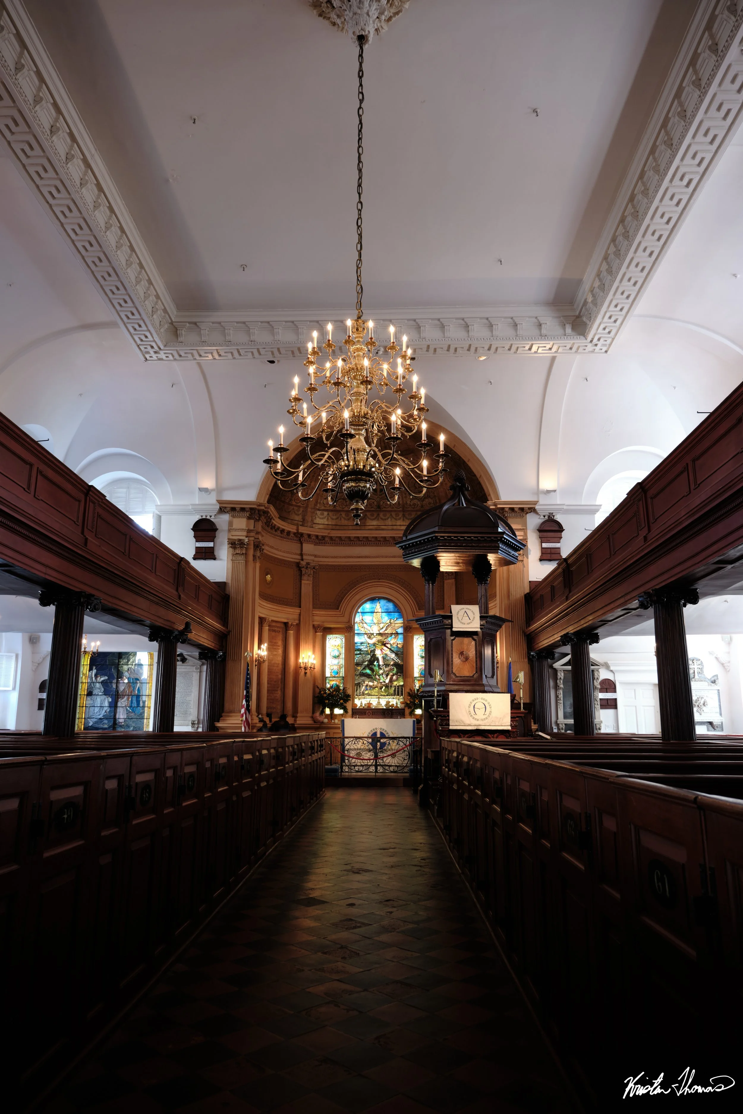 Interior view of a church with wooden pews, a chandelier hanging from the ceiling, stained glass windows, and an altar at the front.