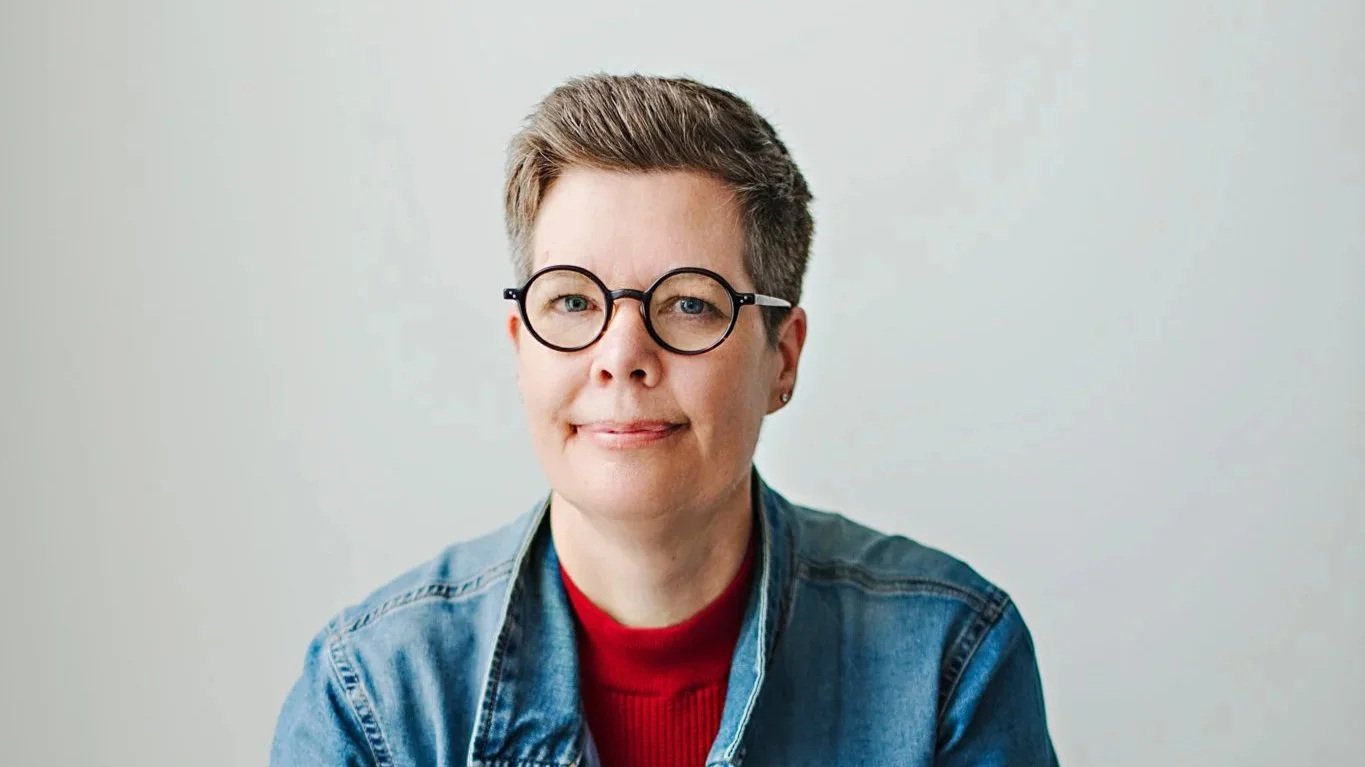 A woman with short hair, glasses, and a small earring, wearing a red shirt and denim jacket, against a plain light background.