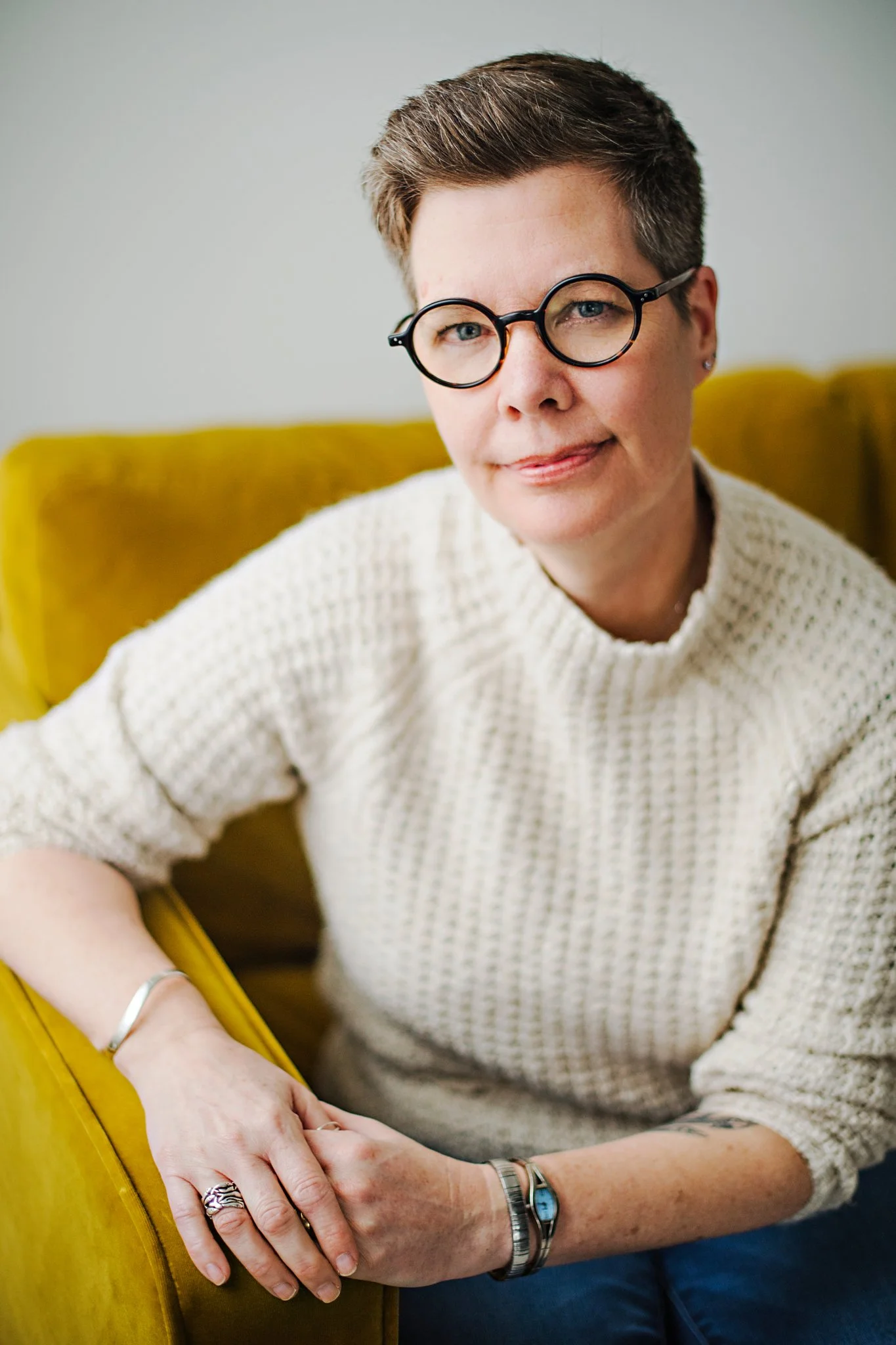 Woman with short brown hair, black glasses, wearing a cream knitted sweater, sitting on a yellow couch, looking at the camera.