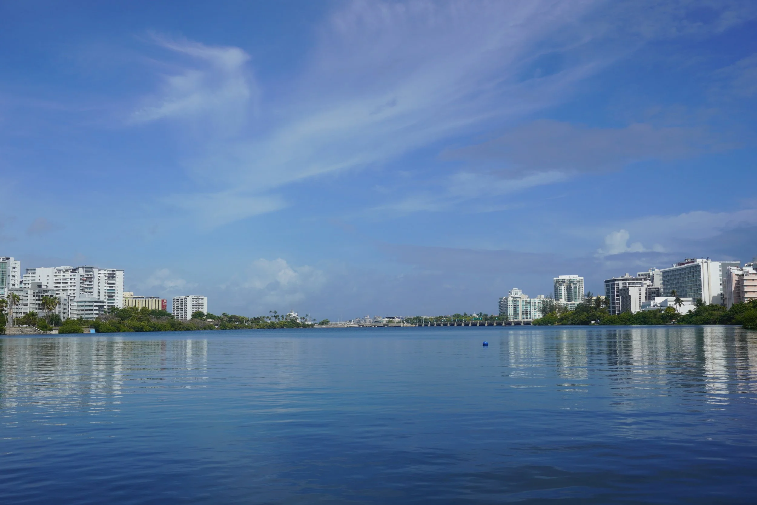 A city skyline with white high-rise buildings along a water body under a blue sky with scattered clouds.