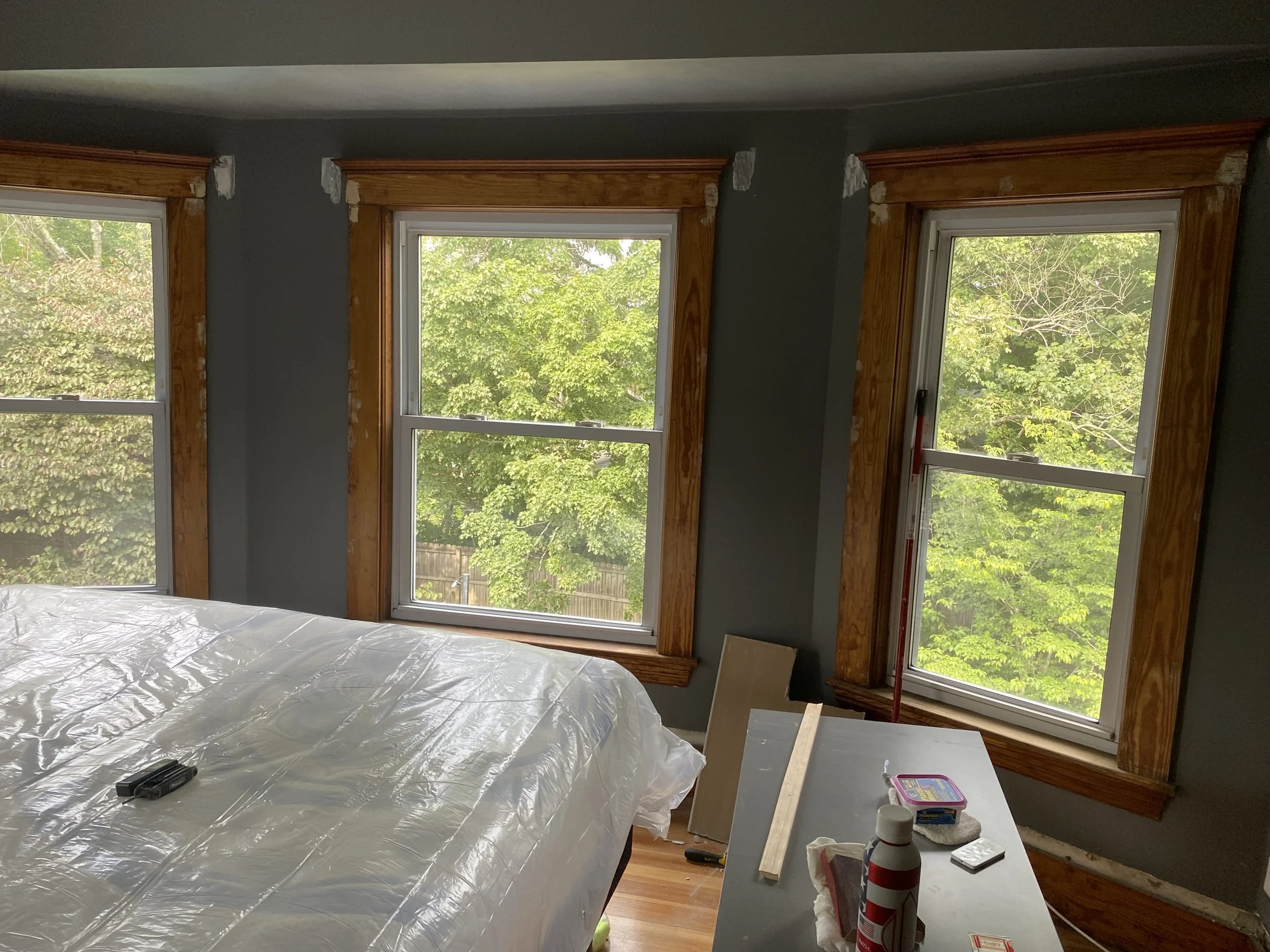 Interior of a room with three large windows, partly covered with wooden trim, with a bed in the foreground and a table with construction supplies.
