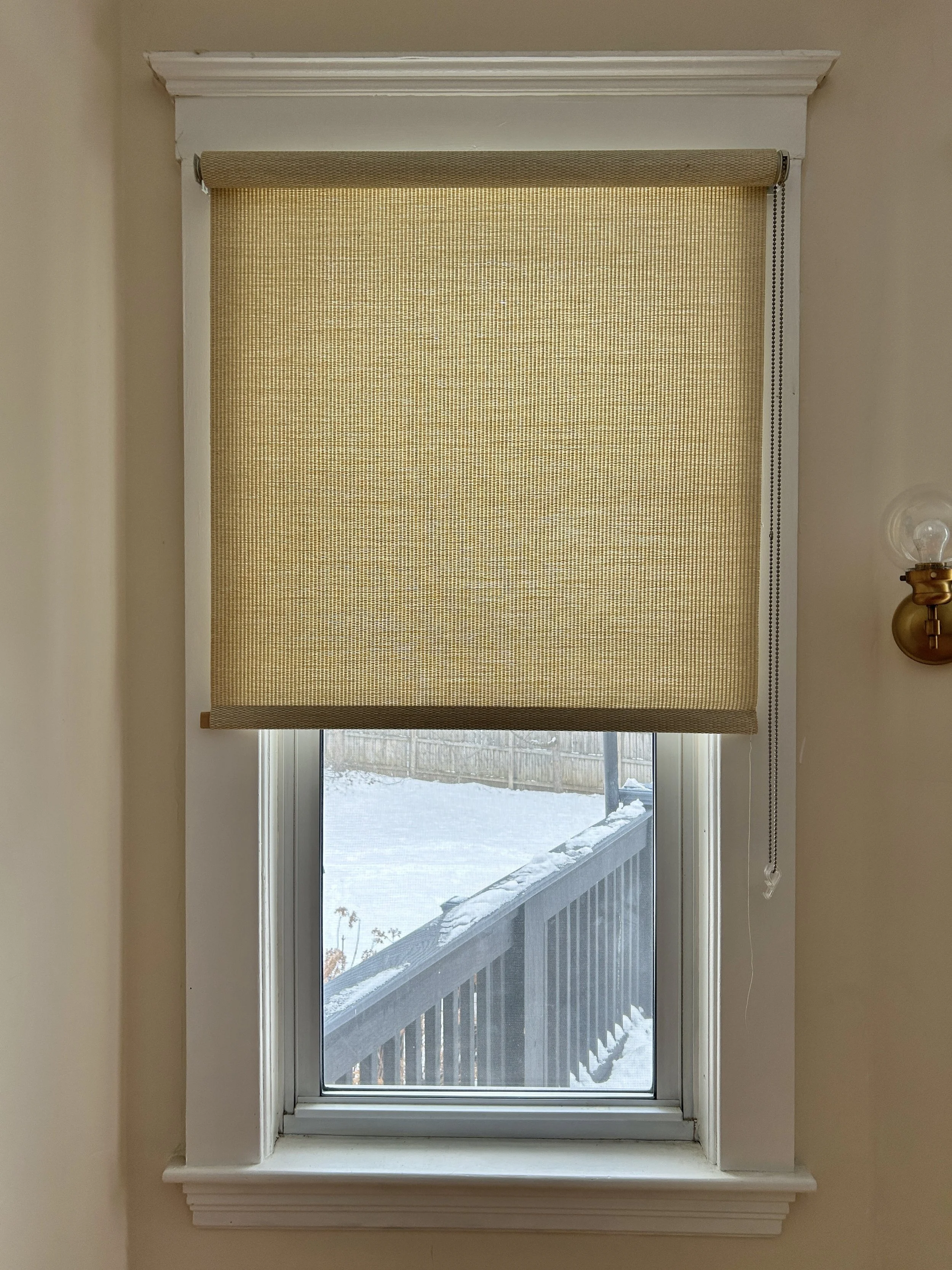 A window with a closed beige roller shade, partially revealing a snowy outdoor scene and a wooden deck railing.
