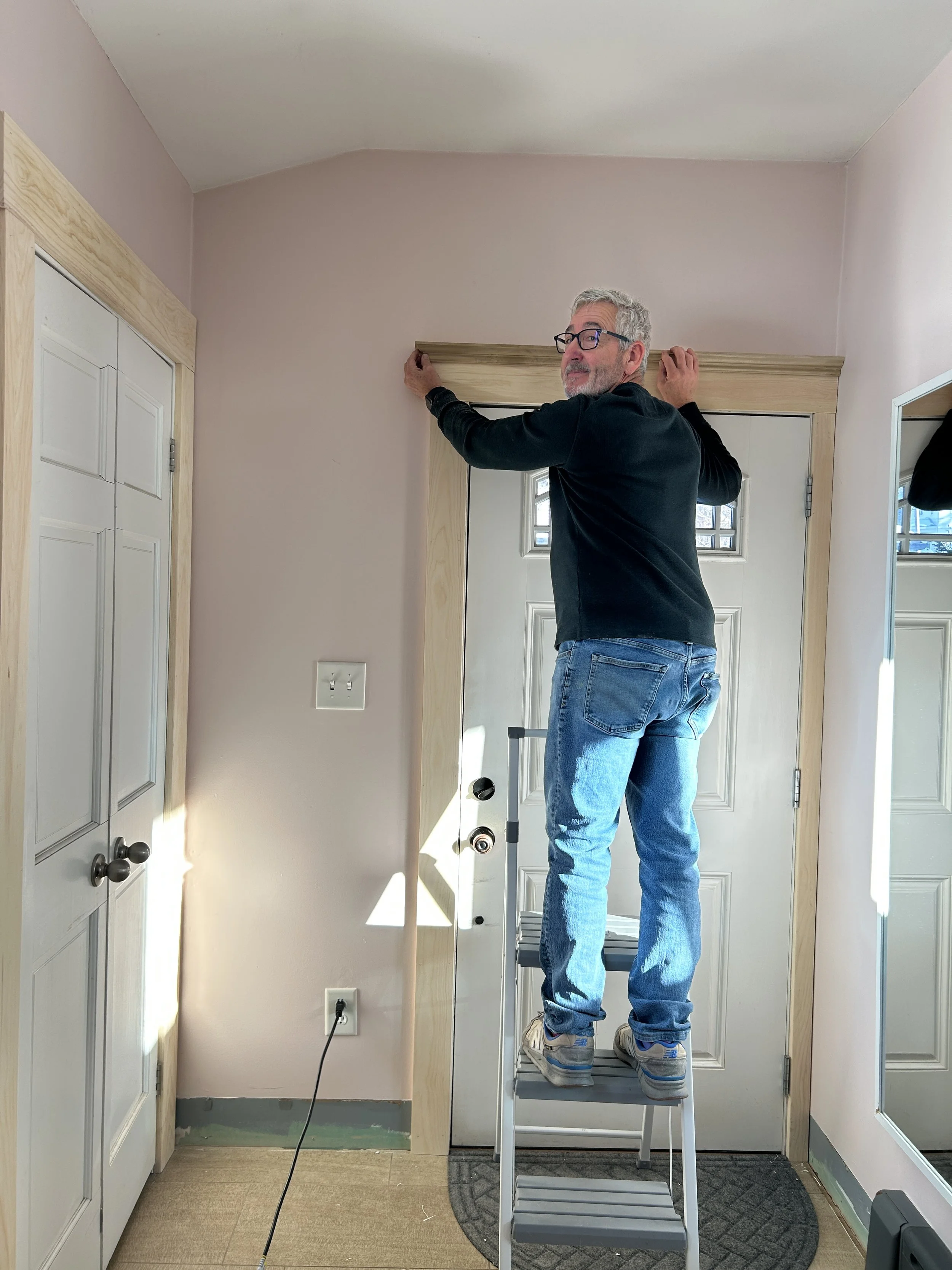 A man standing on a ladder installing a decorative wooden trim above a door in a room.