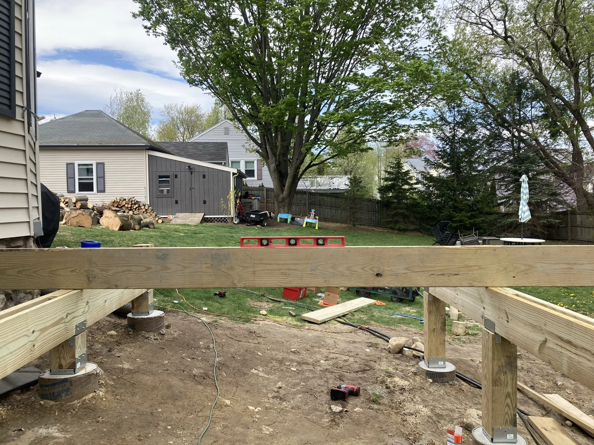 A backyard under construction with wooden railings being installed, construction tools on the ground, a level, and a mound of dirt. The yard has grass, trees, and patio furniture with an umbrella and a fire pit.